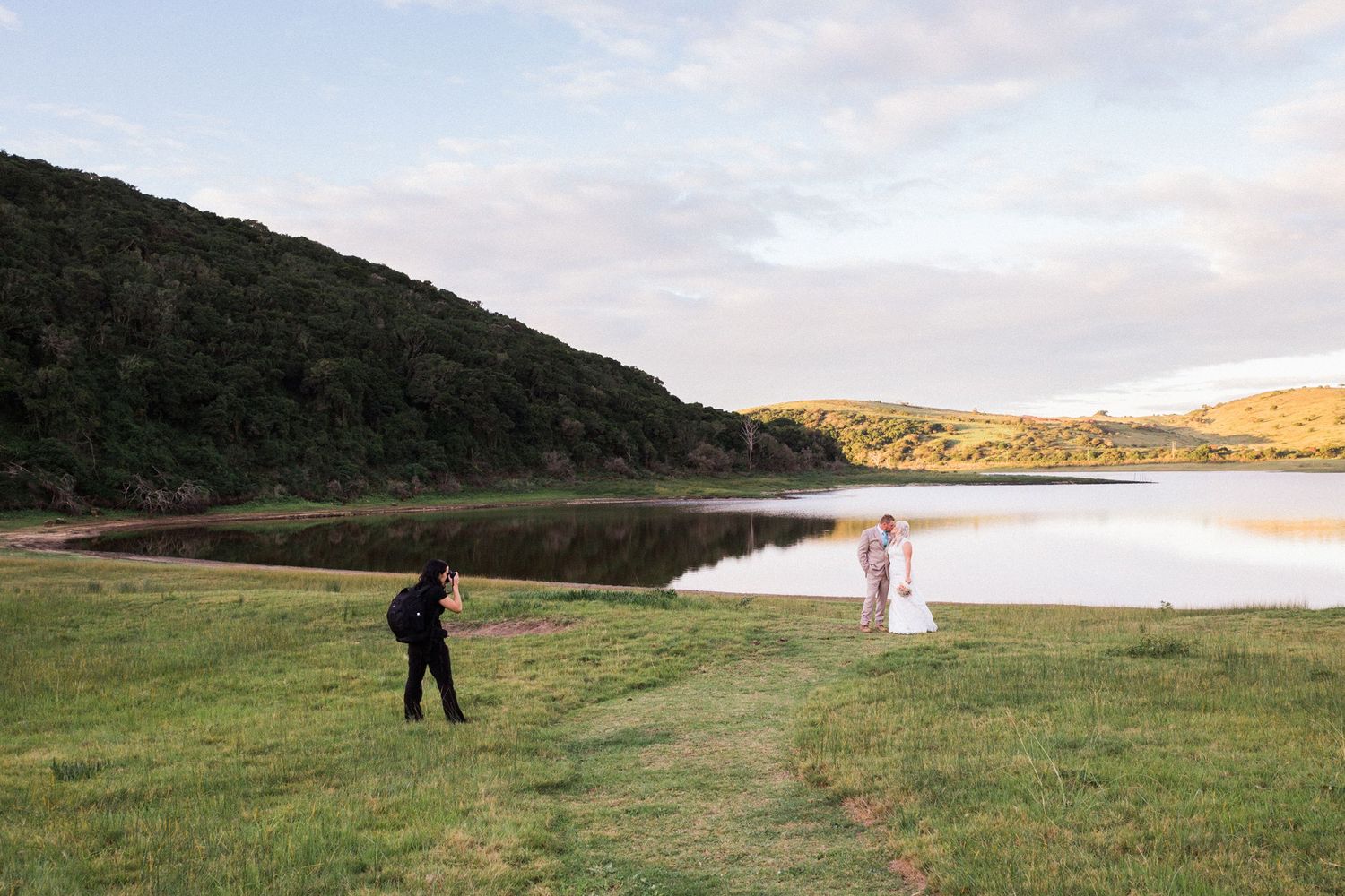 A person stands in a green field overlooking a scenic lake at sunset with rolling hills in the background.
