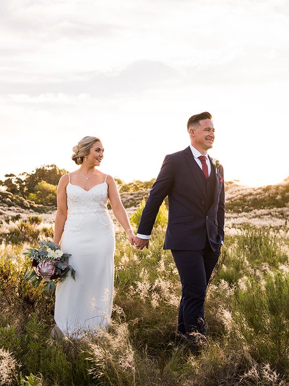 Bride and groom hold hands walking through golden field at sunset, she in white lace gown, he in navy suit.