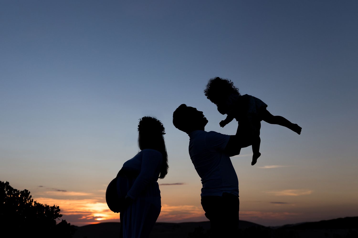 Silhouetted figures stand against a colorful sunset sky in a romantic pose at the top of a hill.