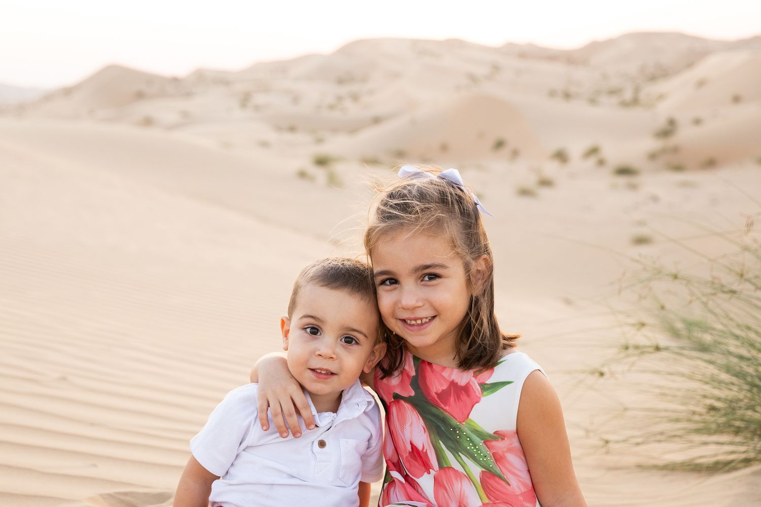 Two children sit close together and play in beige sand dunes on a bright sunny day.