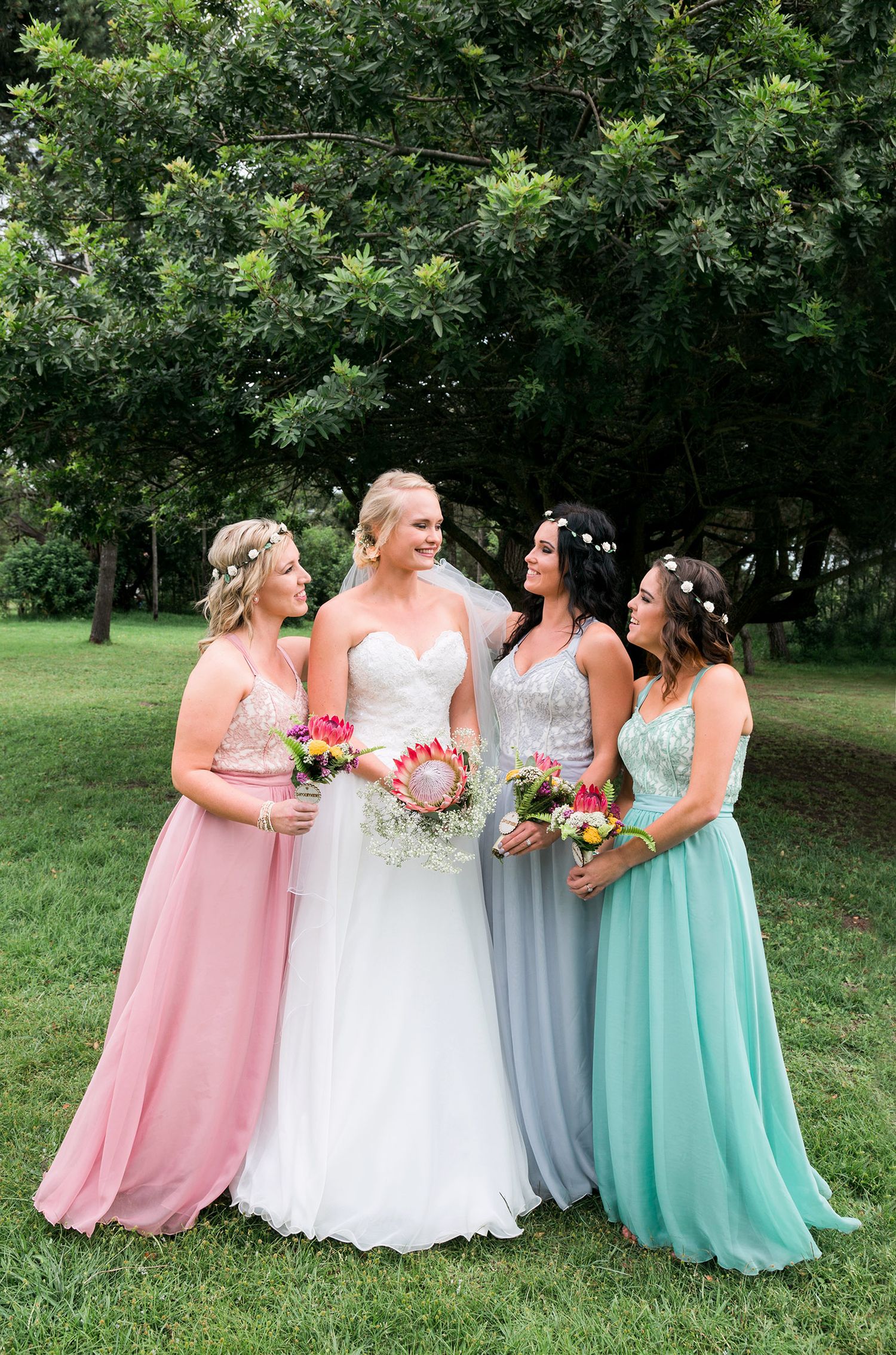 A laughing group in pastel bridesmaid dresses and white wedding gown gather together on a grassy lawn.