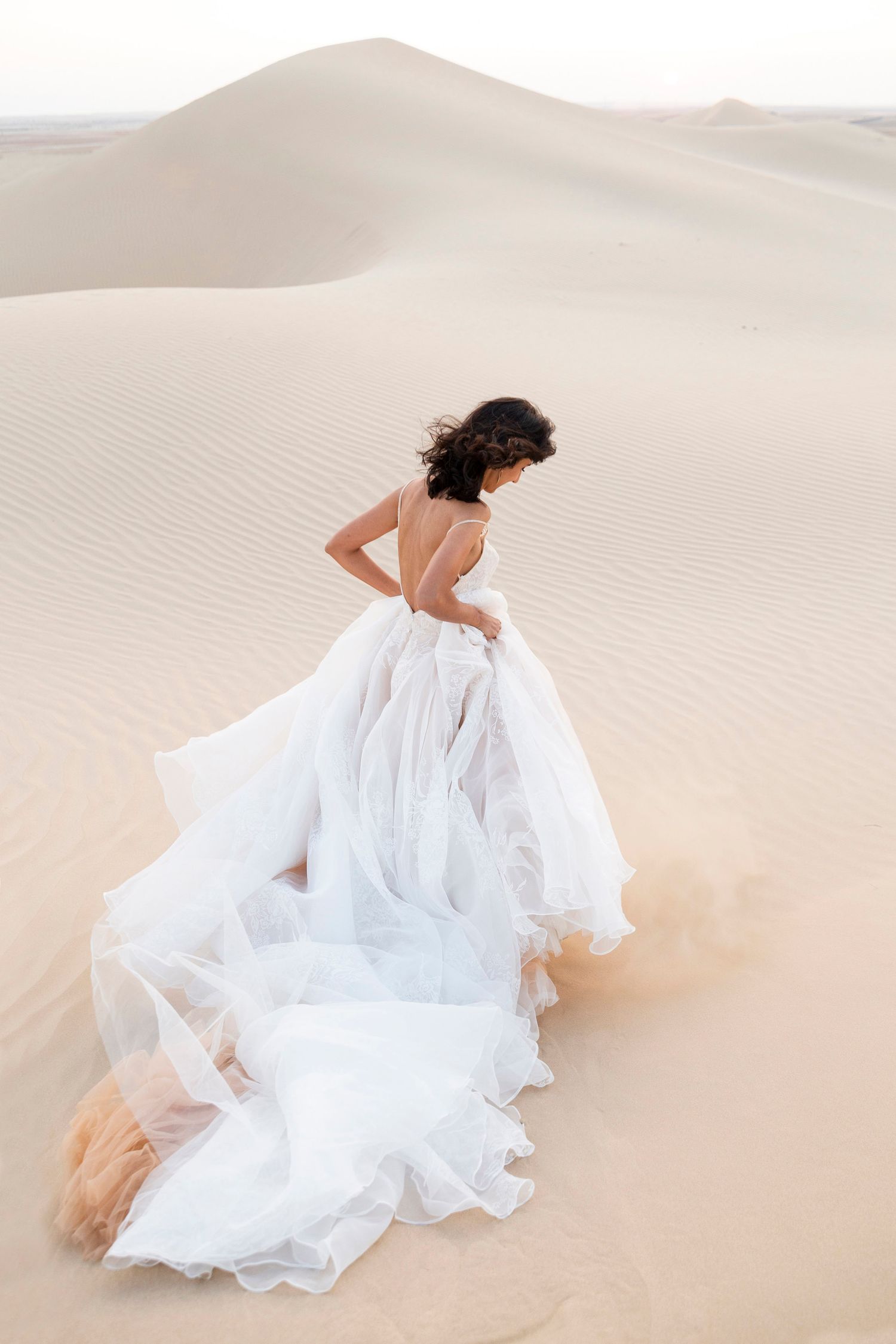 A figure in a flowing white dress reclines gracefully on pristine sand dunes beneath a bright desert sky.