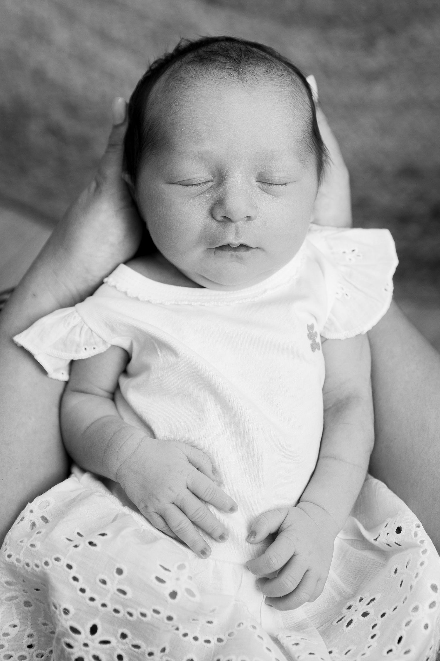 A peaceful sleeping newborn baby wearing white eyelet lace dress captured in black and white portrait photography.