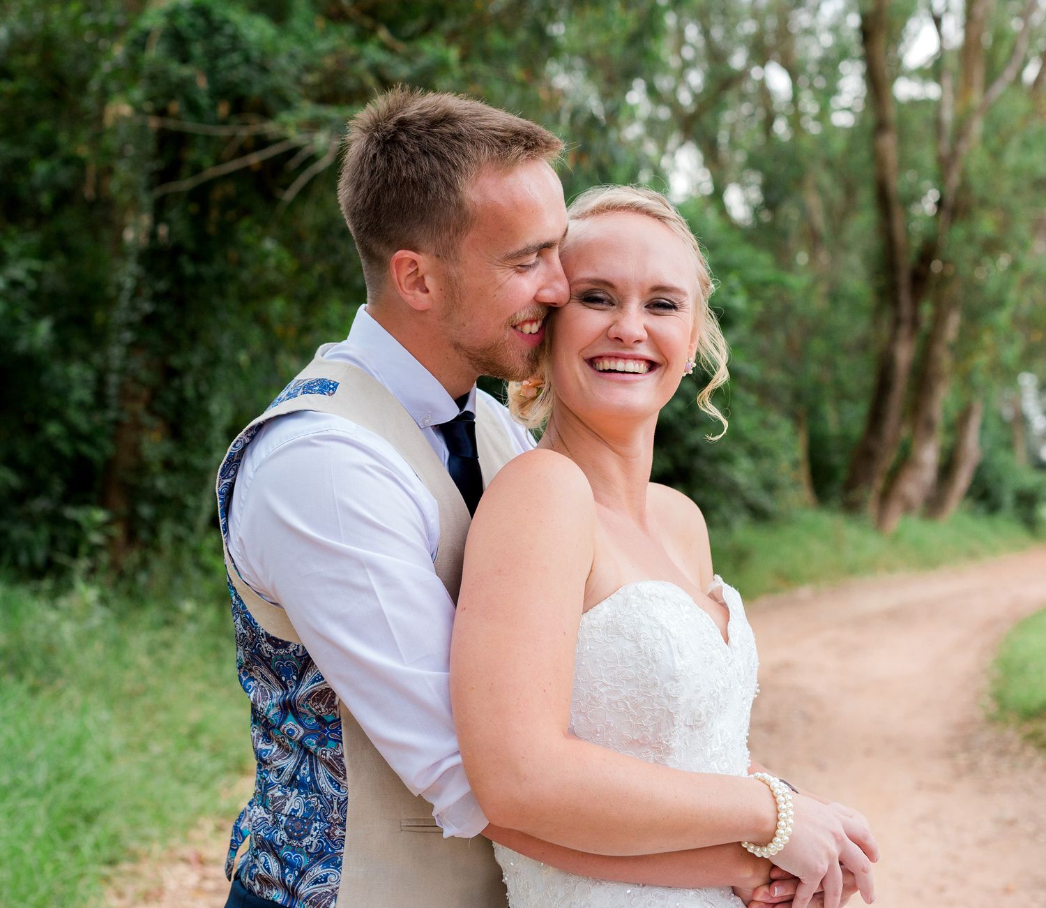 Happy couple shares an intimate moment together laughing and embracing on a tree-lined dirt path on their wedding day.
