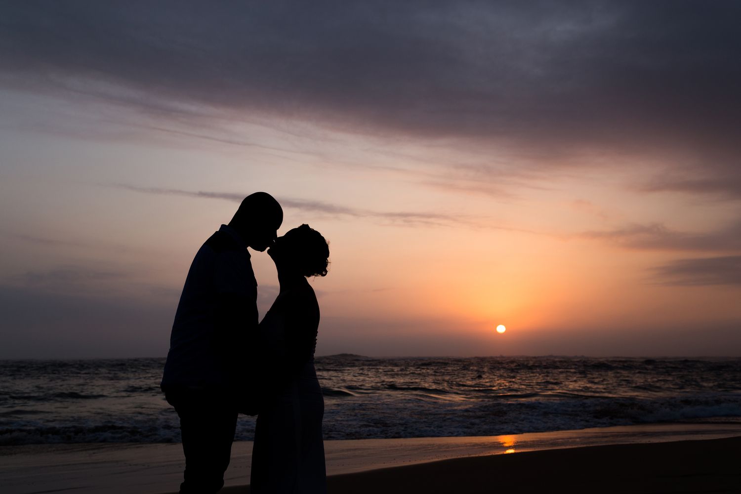Silhouettes of two figures on a beach at sunset with dramatic cloudy sky.