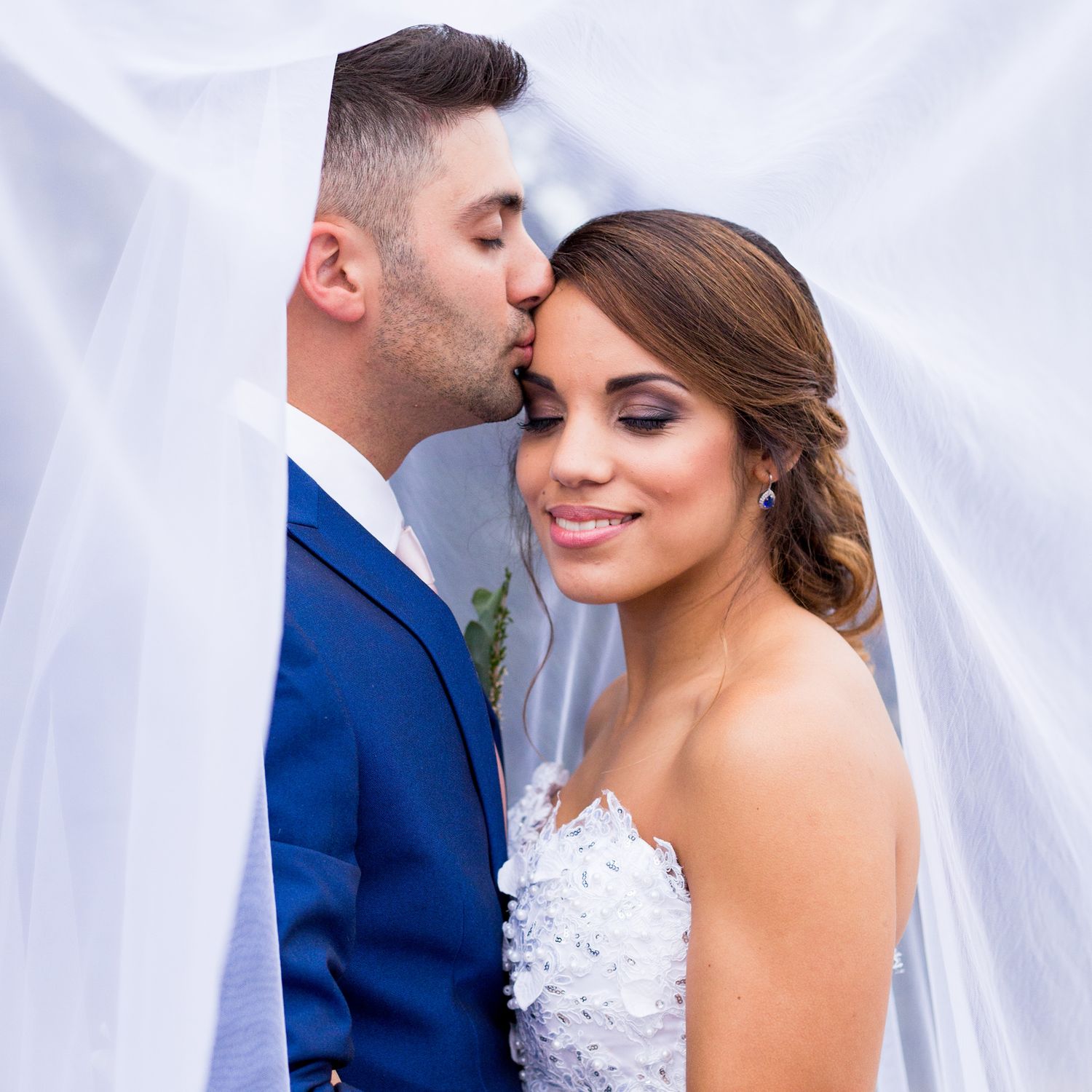 A romantic wedding couple shares an intimate moment in front of a white curtained background while dressed in formal attire.