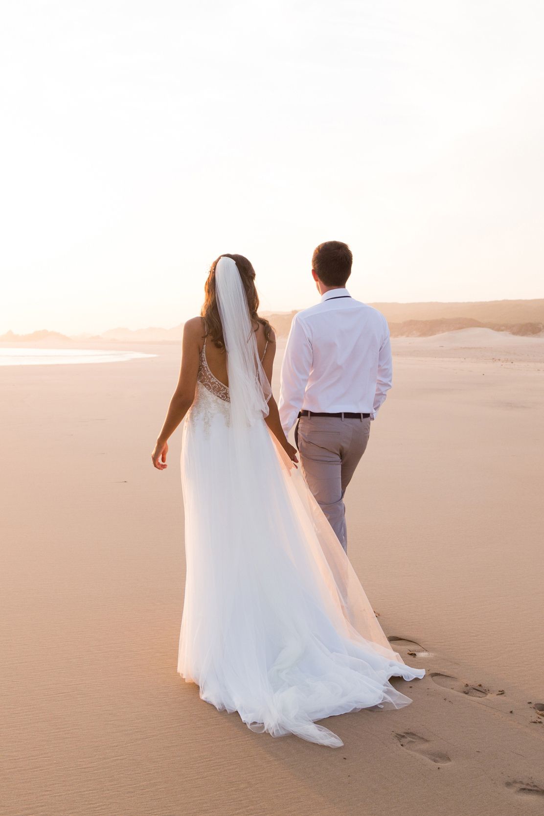 Newlywed couple walking together at sunset leaving footprints in sand dunes during romantic desert photoshoot.