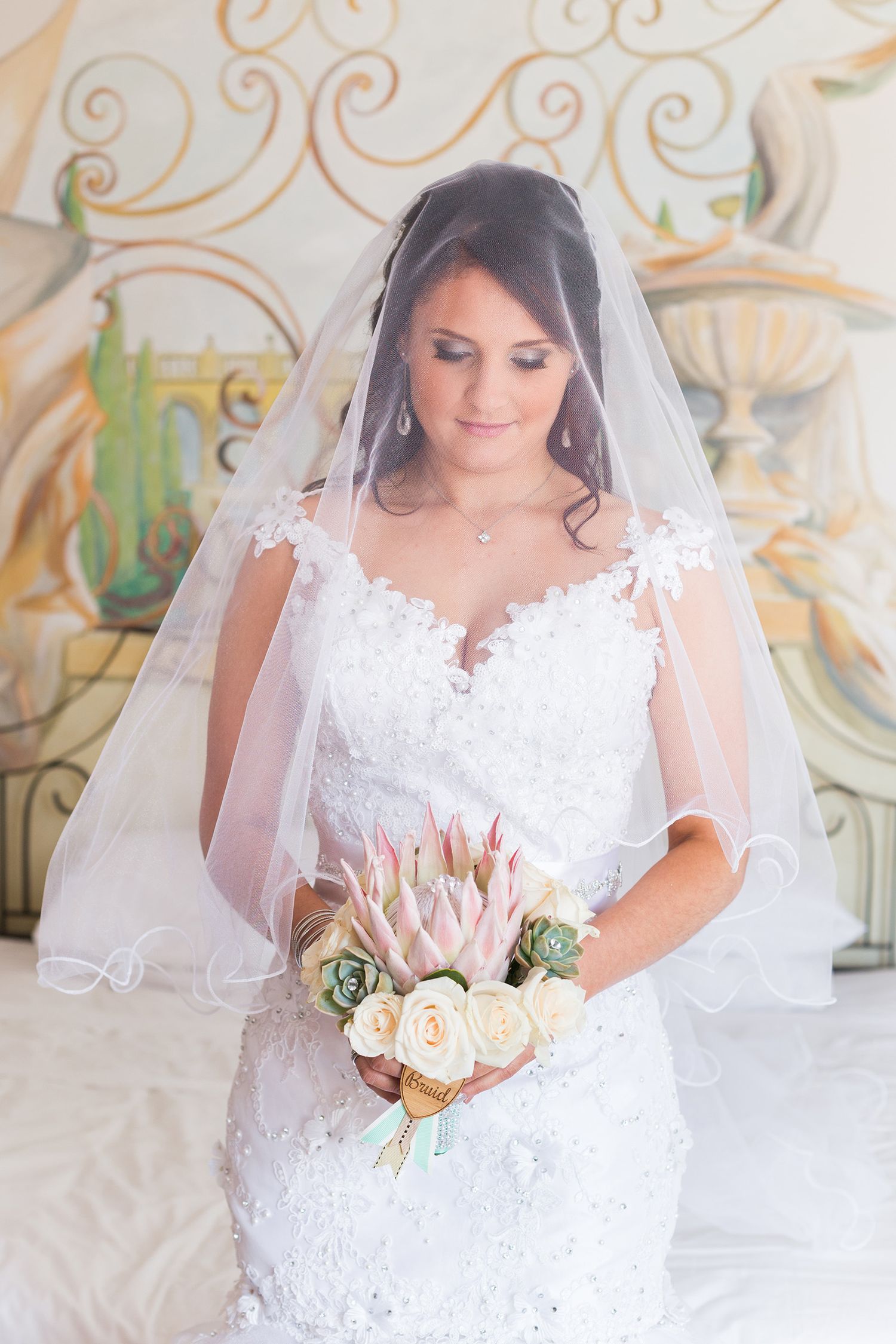 A stunning bride in a white lace wedding gown and veil poses elegantly with a peach and blush rose bouquet.