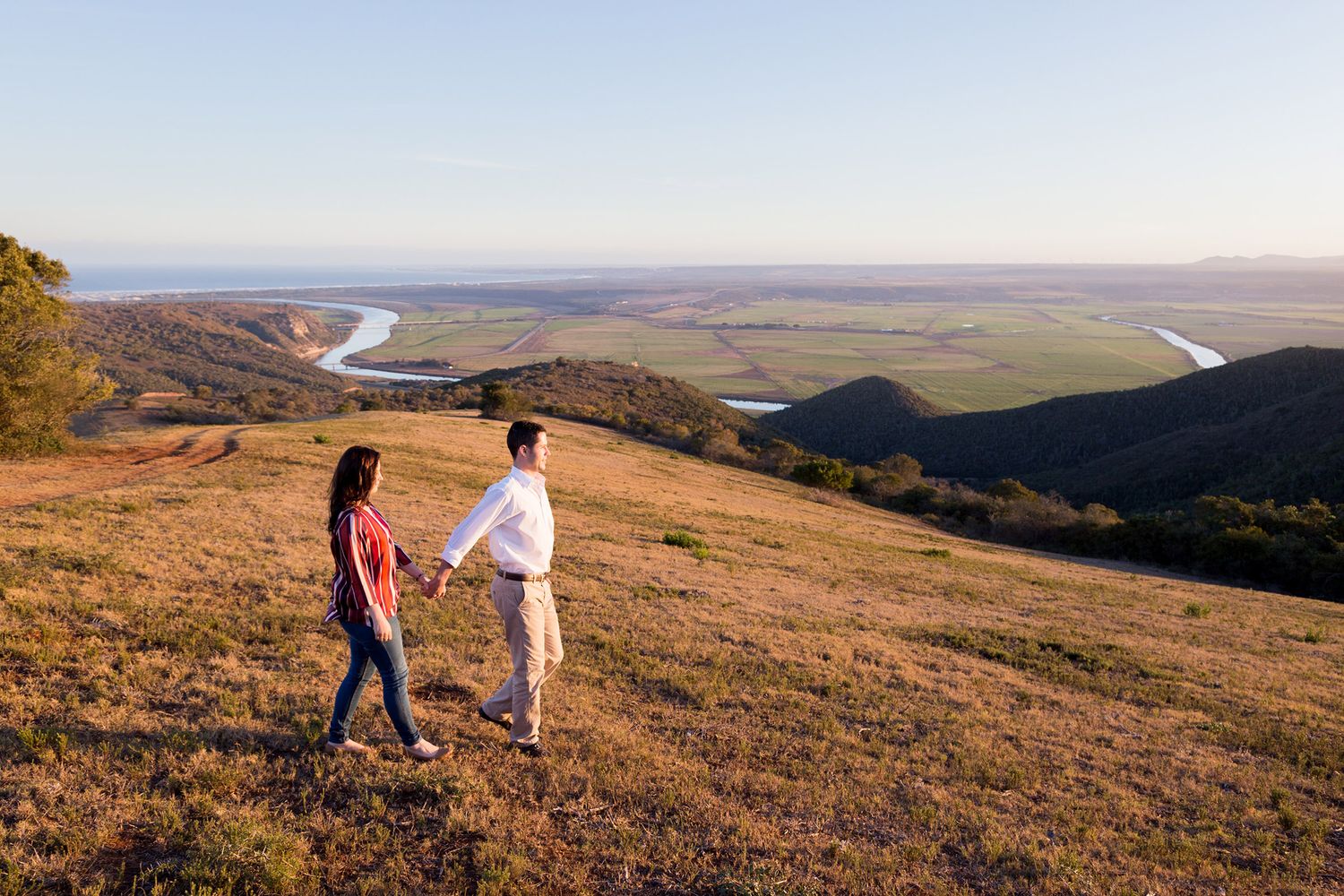 Two people walk together along a scenic hillside path overlooking mountains and valleys at sunset.