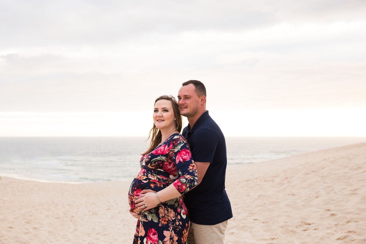 Woman in floral dress and partner walk together along a scenic beach at sunset with ocean views.