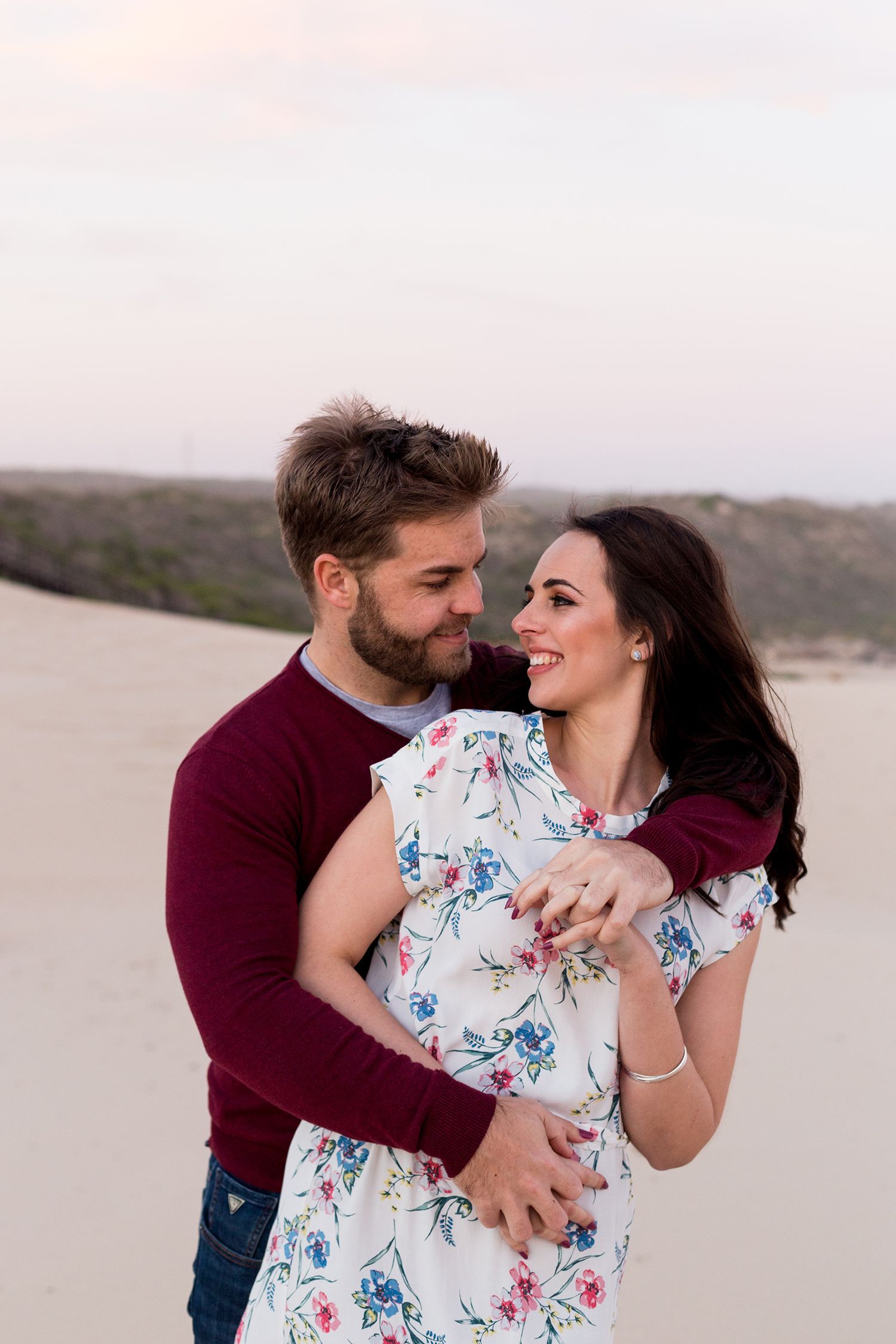 A couple embraces on a sandy beach at sunset wearing burgundy and floral print outfits for a romantic photoshoot.