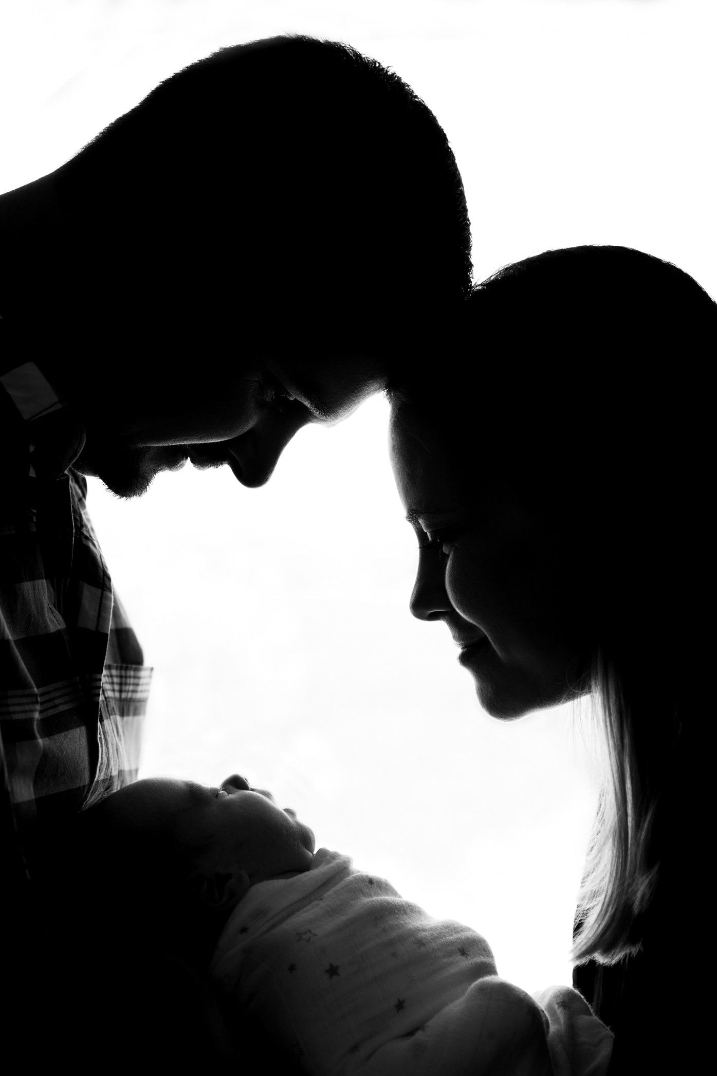 Black and white silhouette photo shows parents lovingly gazing at their newborn baby against white background.