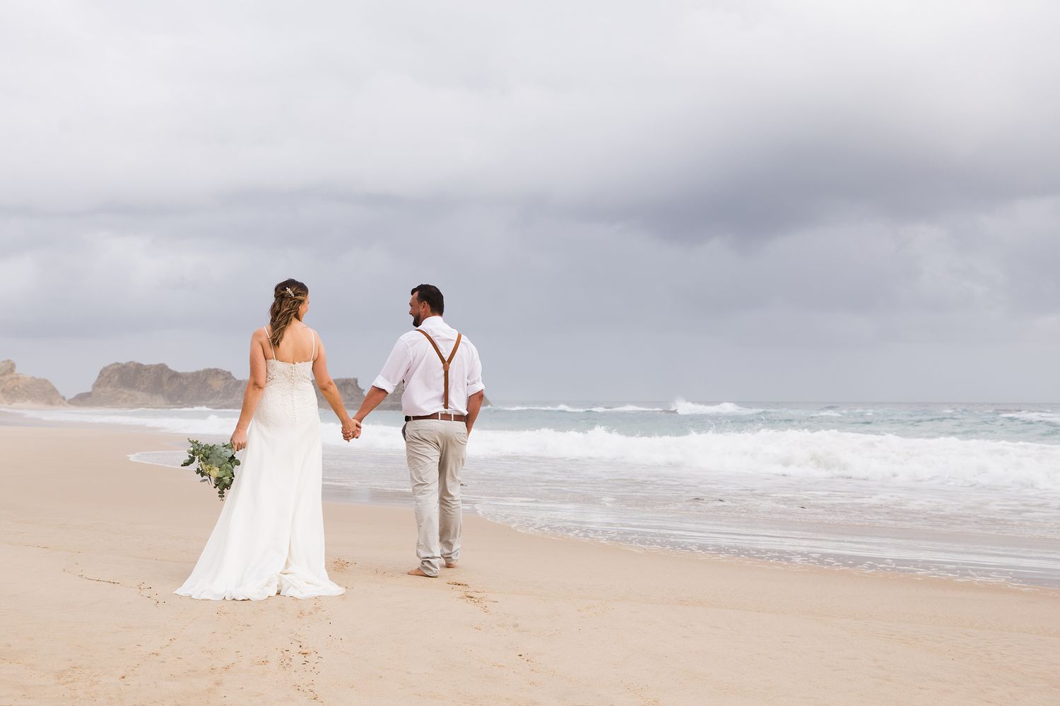 A couple in white attire hold hands while walking together on a scenic beach with waves crashing in the background.