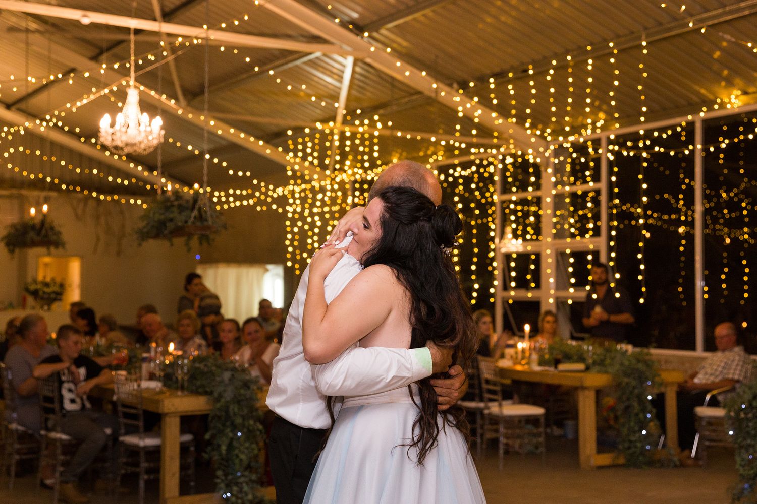 A couple dancing at a wedding reception under warm twinkle lights and string lighting in a rustic indoor venue.