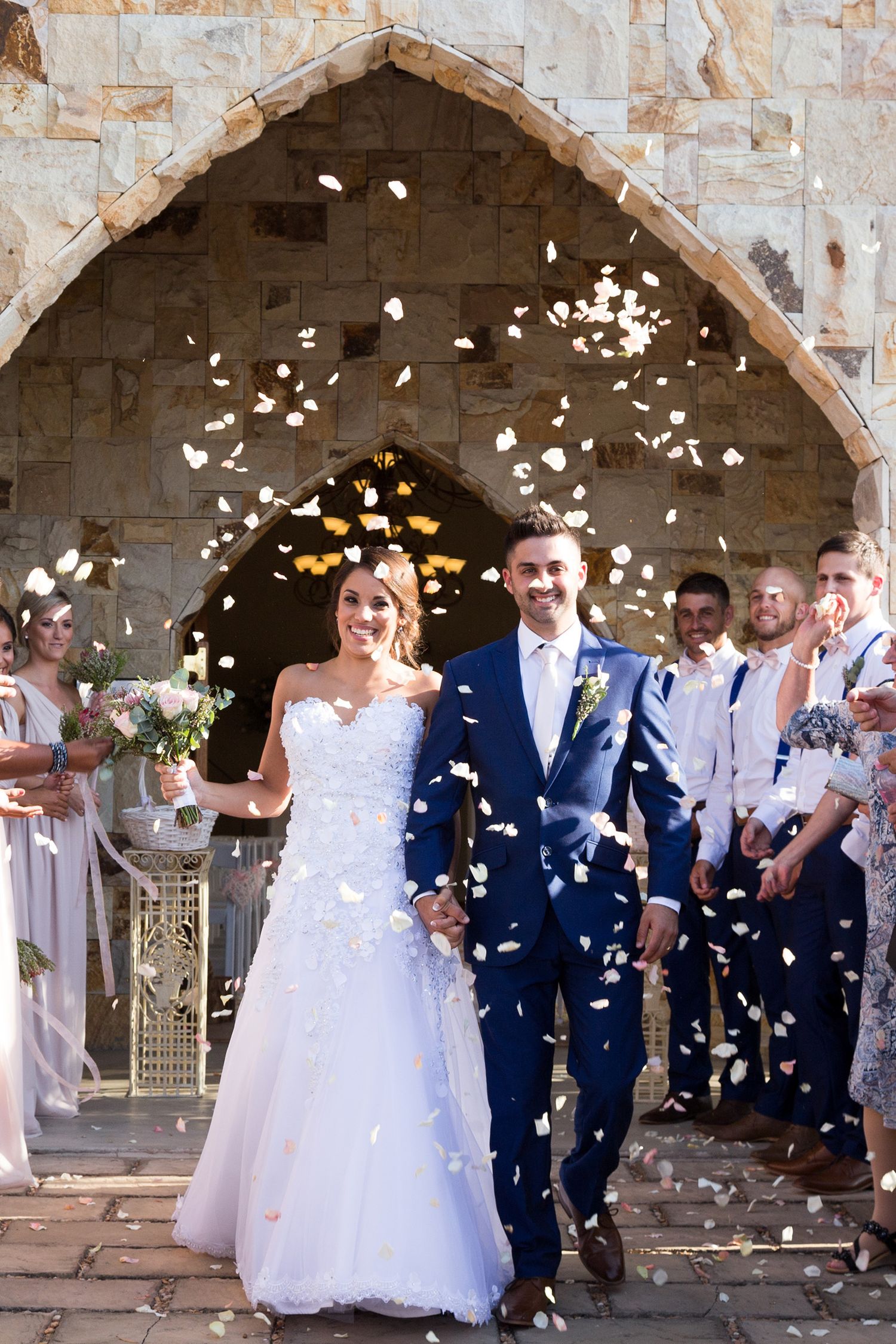 Wedding party celebrates under stone arches as white flower petals shower down on the newlyweds.