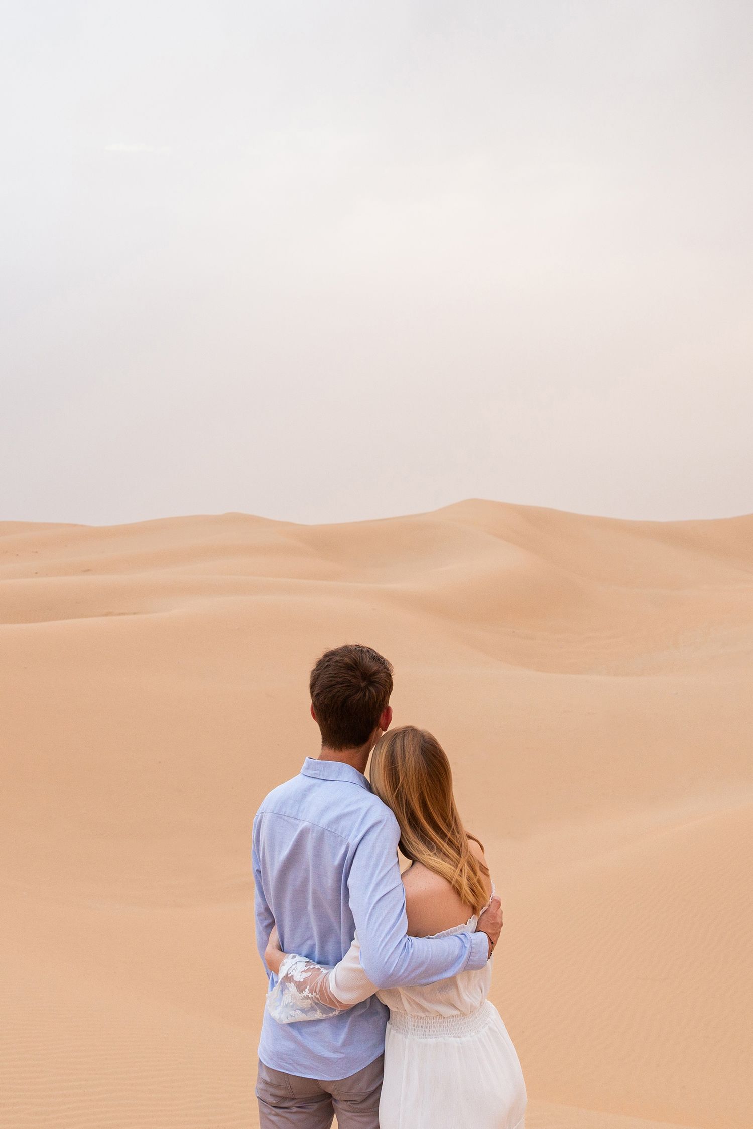 A couple dressed in blue and white embrace while overlooking vast golden sand dunes in the desert.