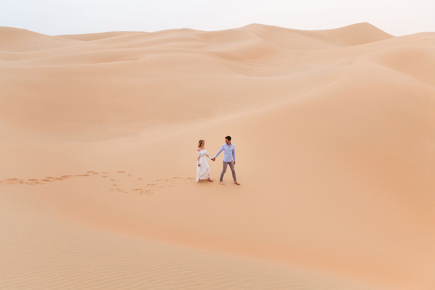 Two figures walking on rolling desert sand dunes against a warm beige sky at sunset.