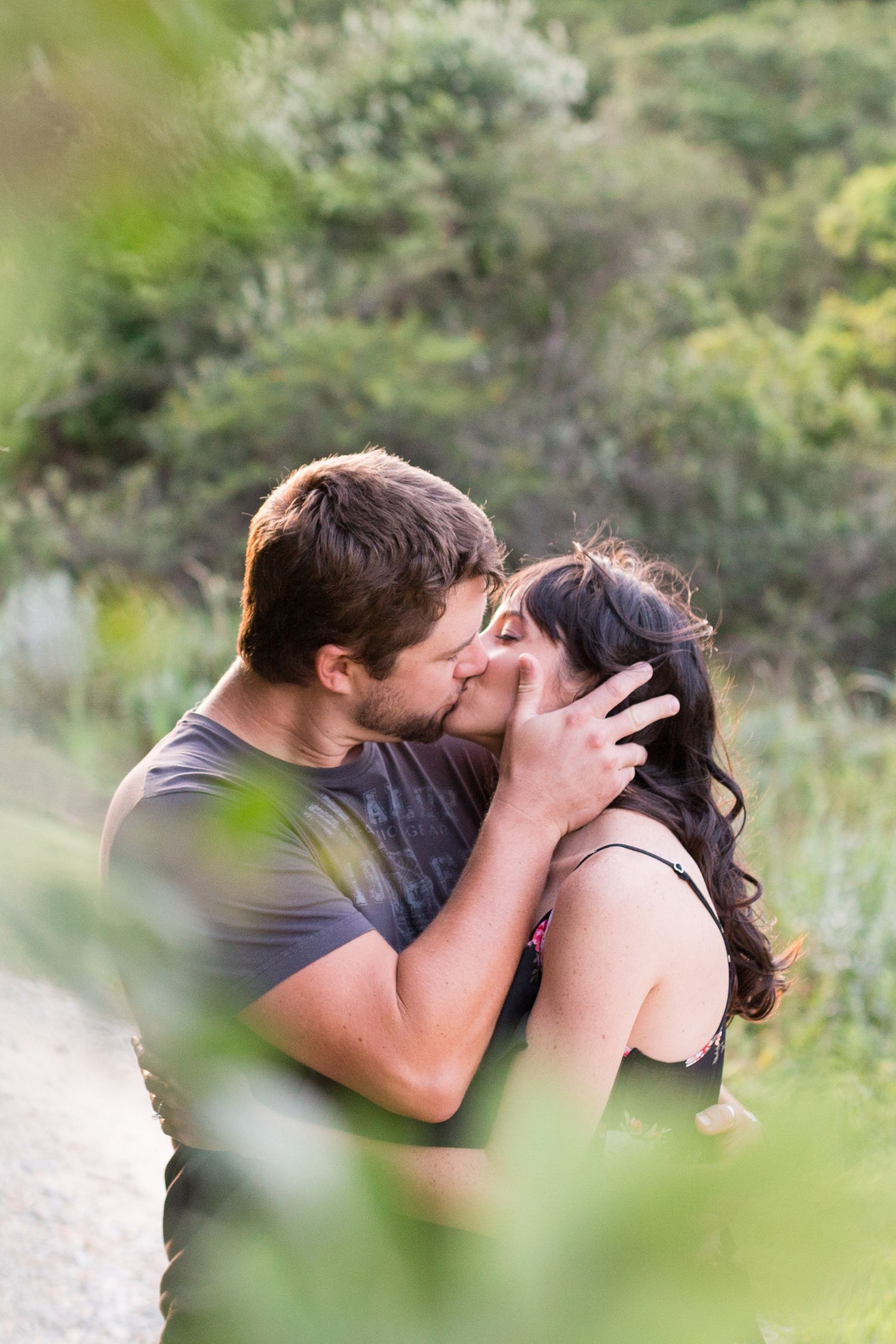 Romantic couple shares intimate kiss while embracing outdoors in nature with blurred greenery in foreground.
