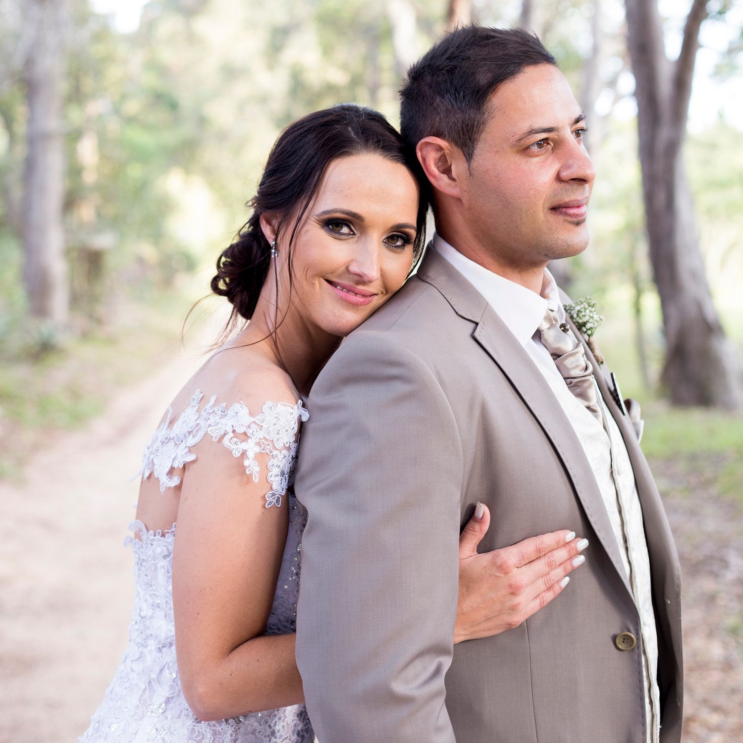 Wedding couple in gray suit and white dress embrace outdoors in soft natural lighting.