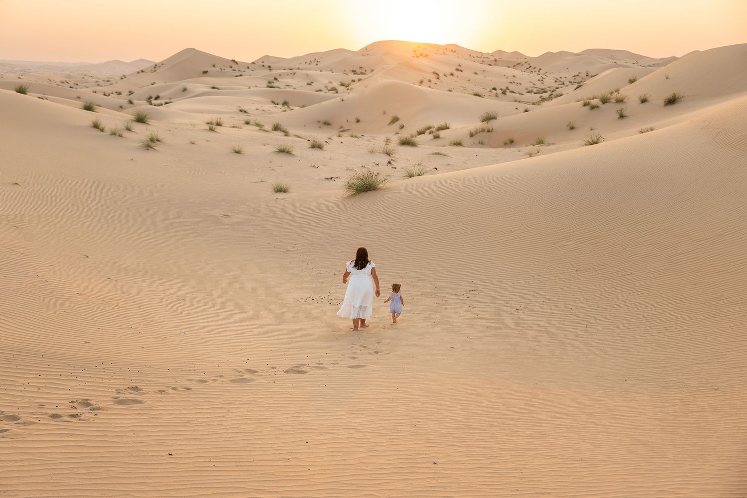 People walk through rolling sand dunes during golden sunset in a vast desert landscape.