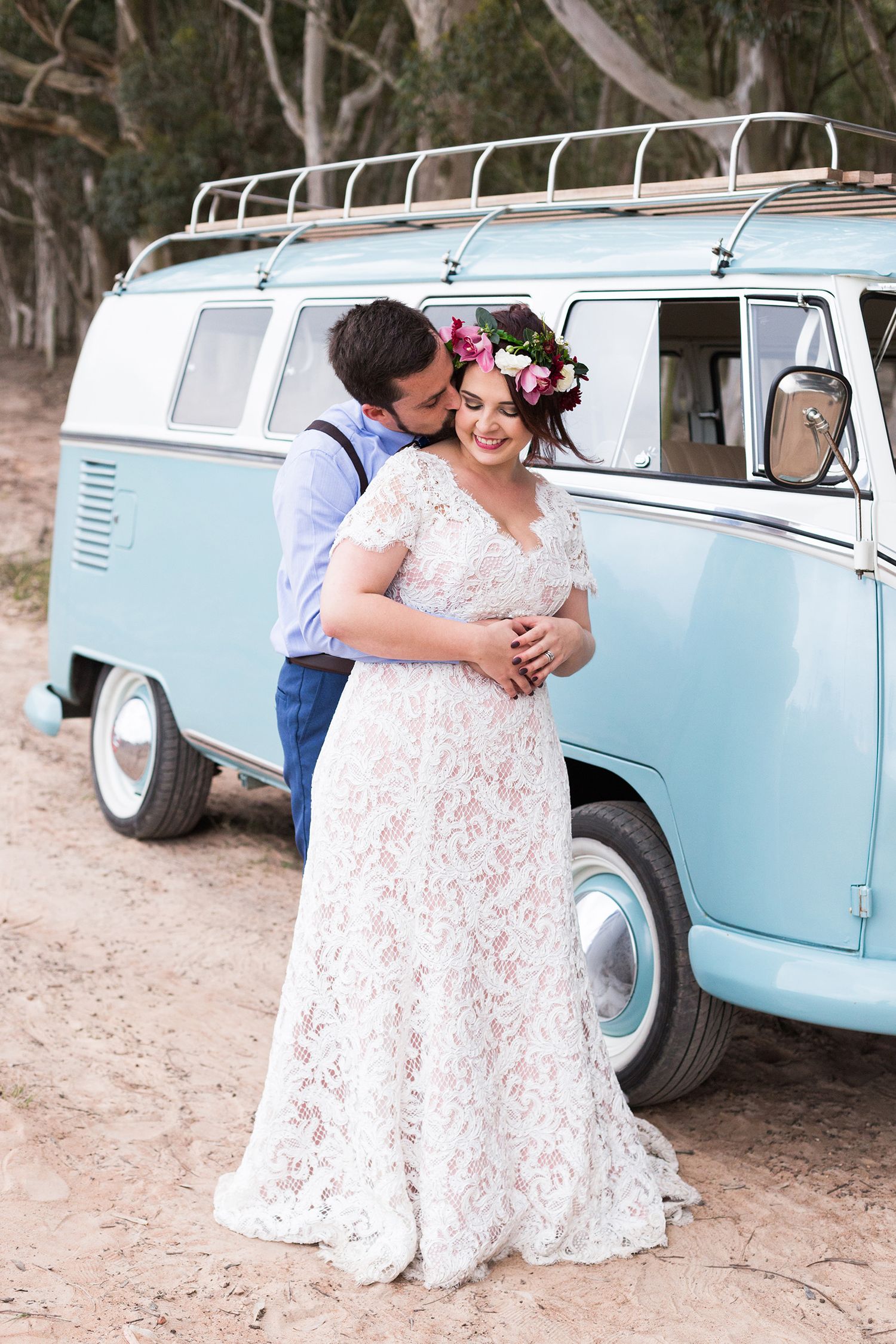 A couple embraces near a light blue vintage Volkswagen bus in a rustic outdoor setting.
