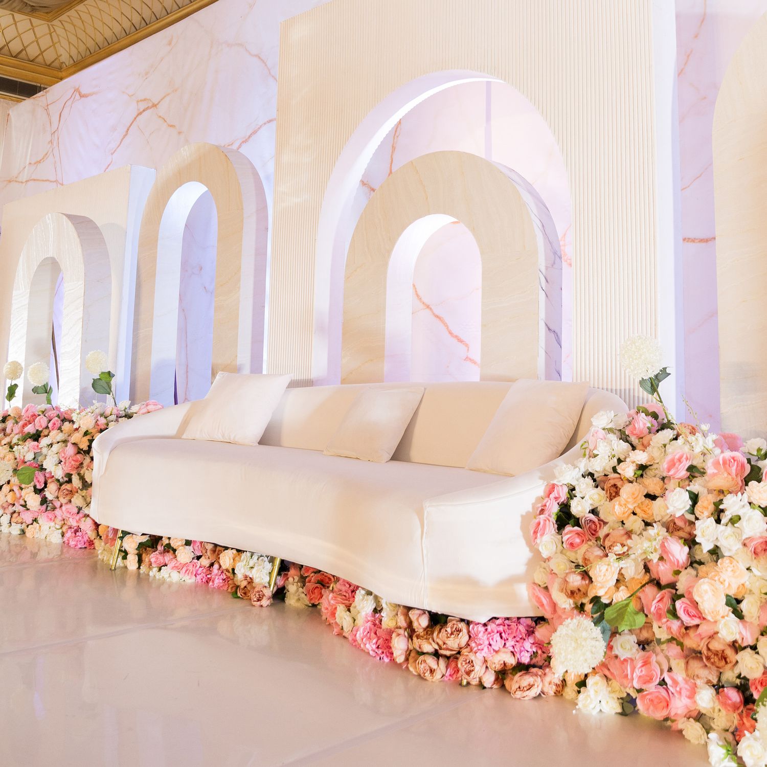 Elegant white curved sofa decorated with cascading pink and peach floral arrangements against arched white walls.