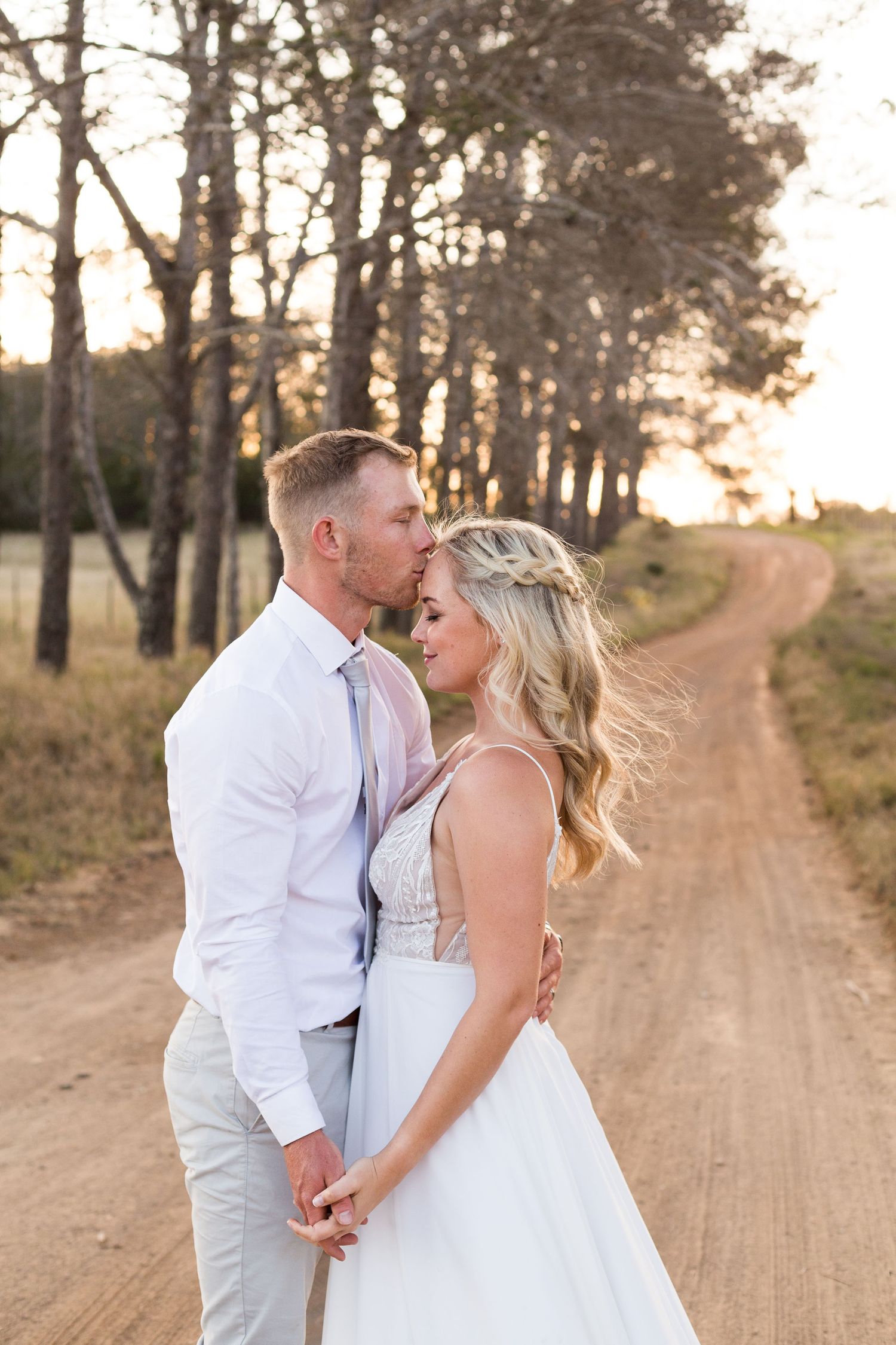 A couple embraces on a dirt road surrounded by tall pine trees at sunset, the bride wearing a flowing white gown.