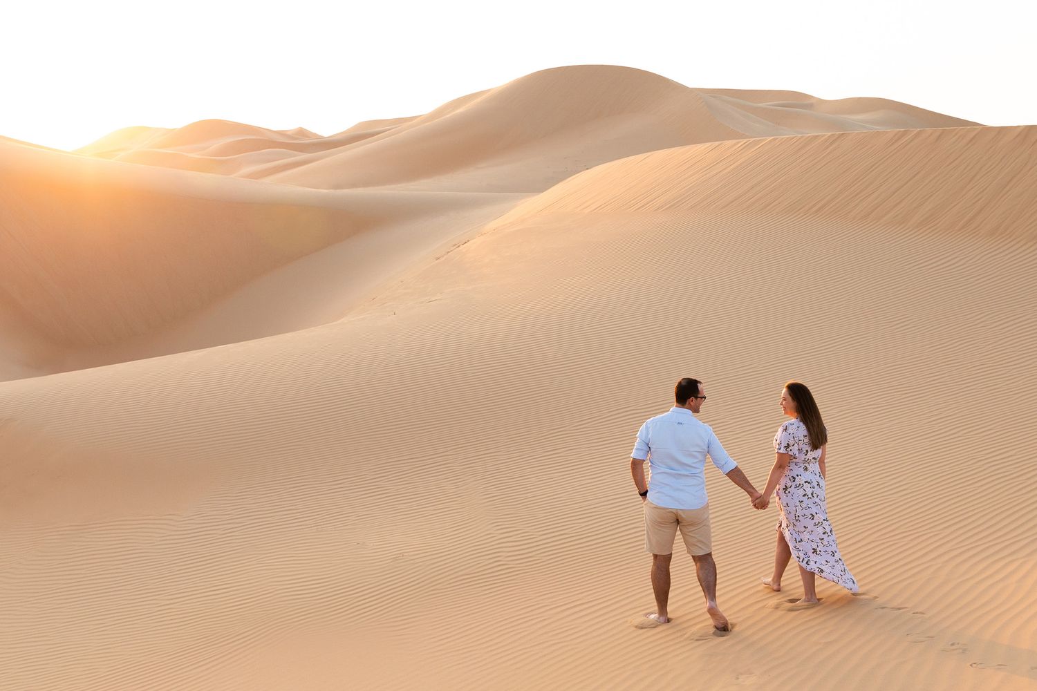 Two people sitting in white attire on golden sand dunes during sunset in a desert landscape.