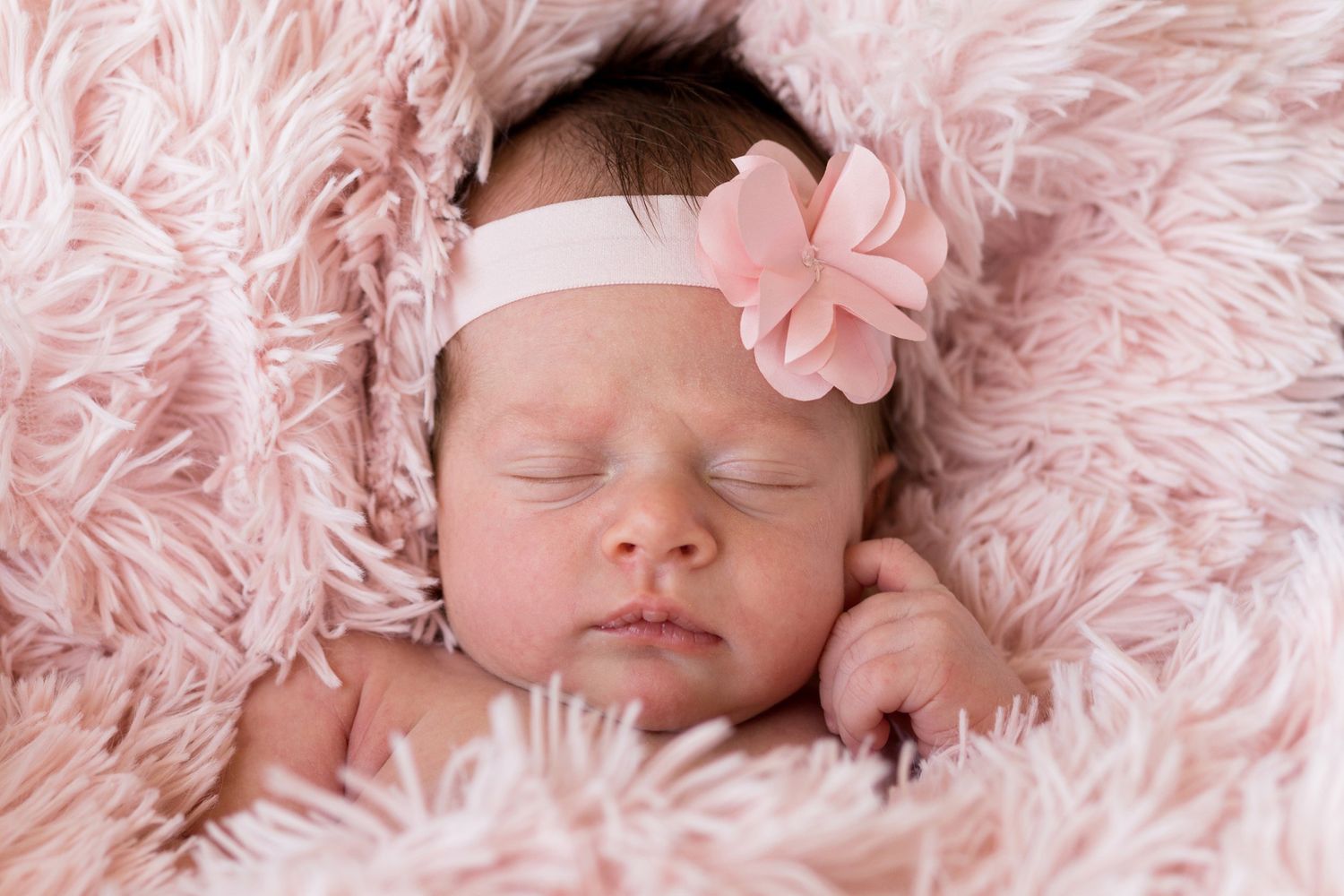 A sleeping newborn baby wrapped in soft pink fur blanket wearing a pink headband with flower.