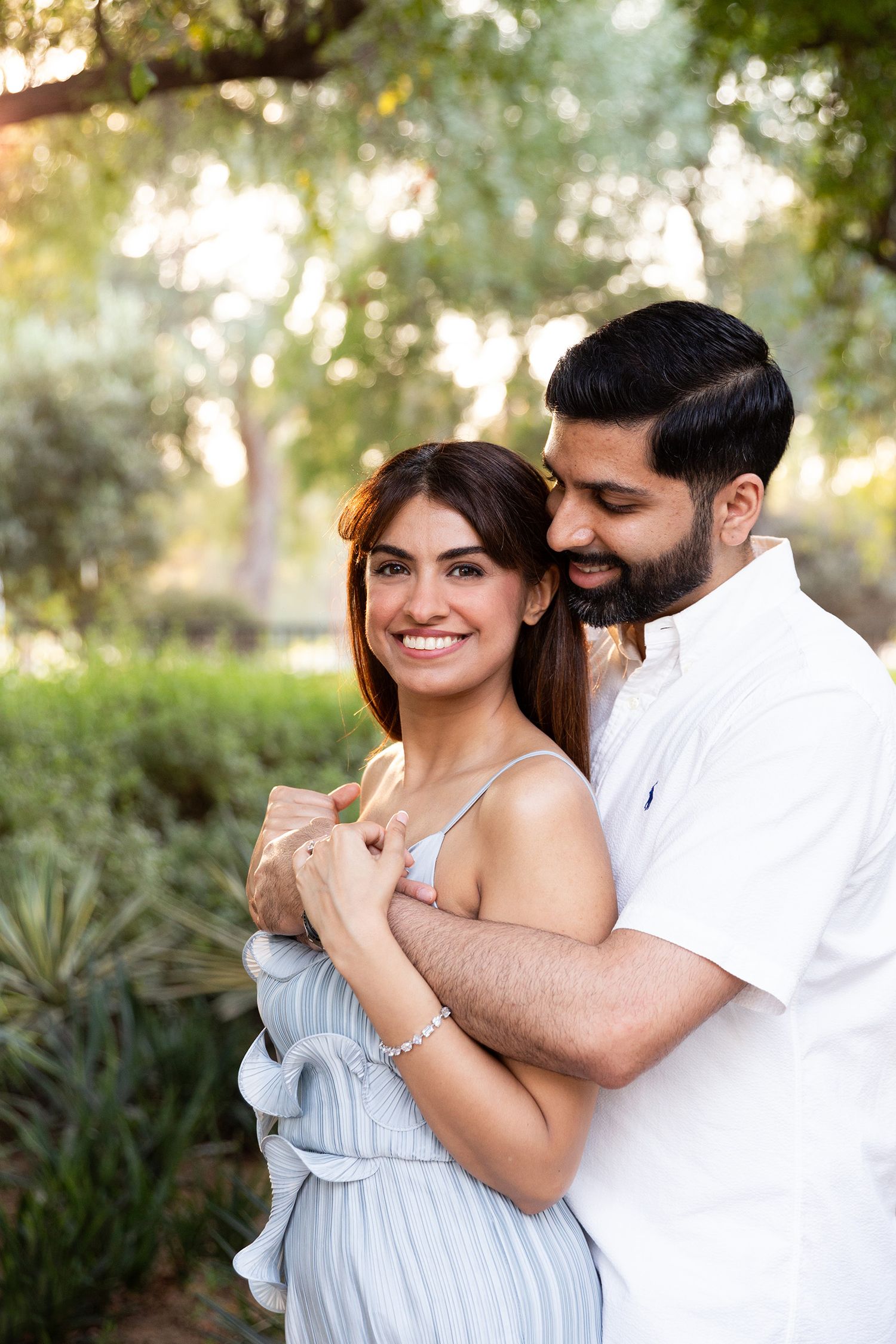 A couple embraces outdoors in a garden setting, with her wearing a light blue dress and him in a white shirt.