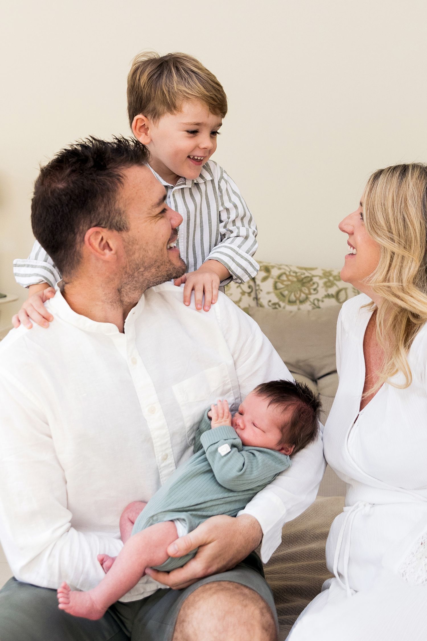 Family lying on bed in white clothing sharing tender moments with their newborn baby in lifestyle photoshoot.