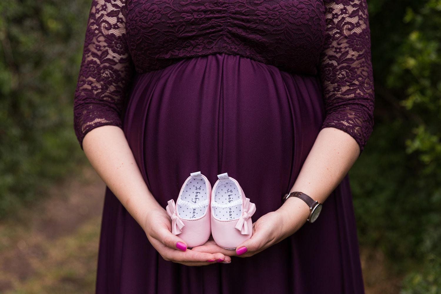 Close-up of tiny white baby shoes being held against a dark plum colored lace dress in an outdoor setting.