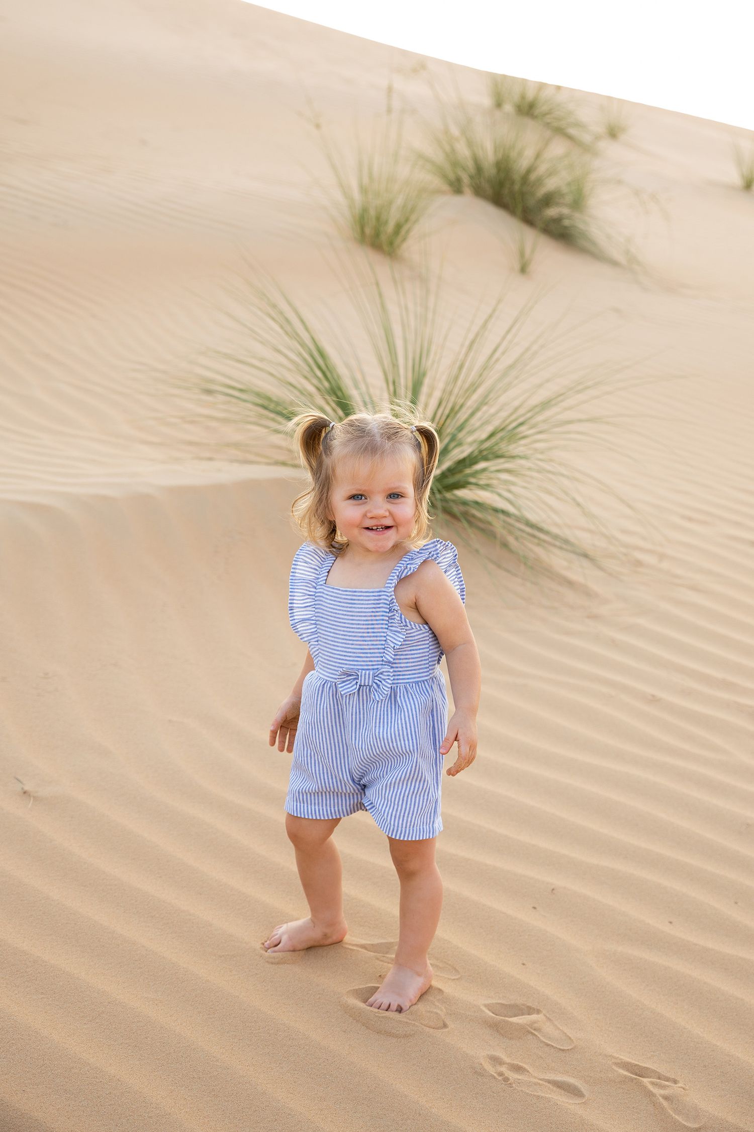 A small child wearing a blue and white striped romper plays barefoot in sandy dunes with scattered beach grass.