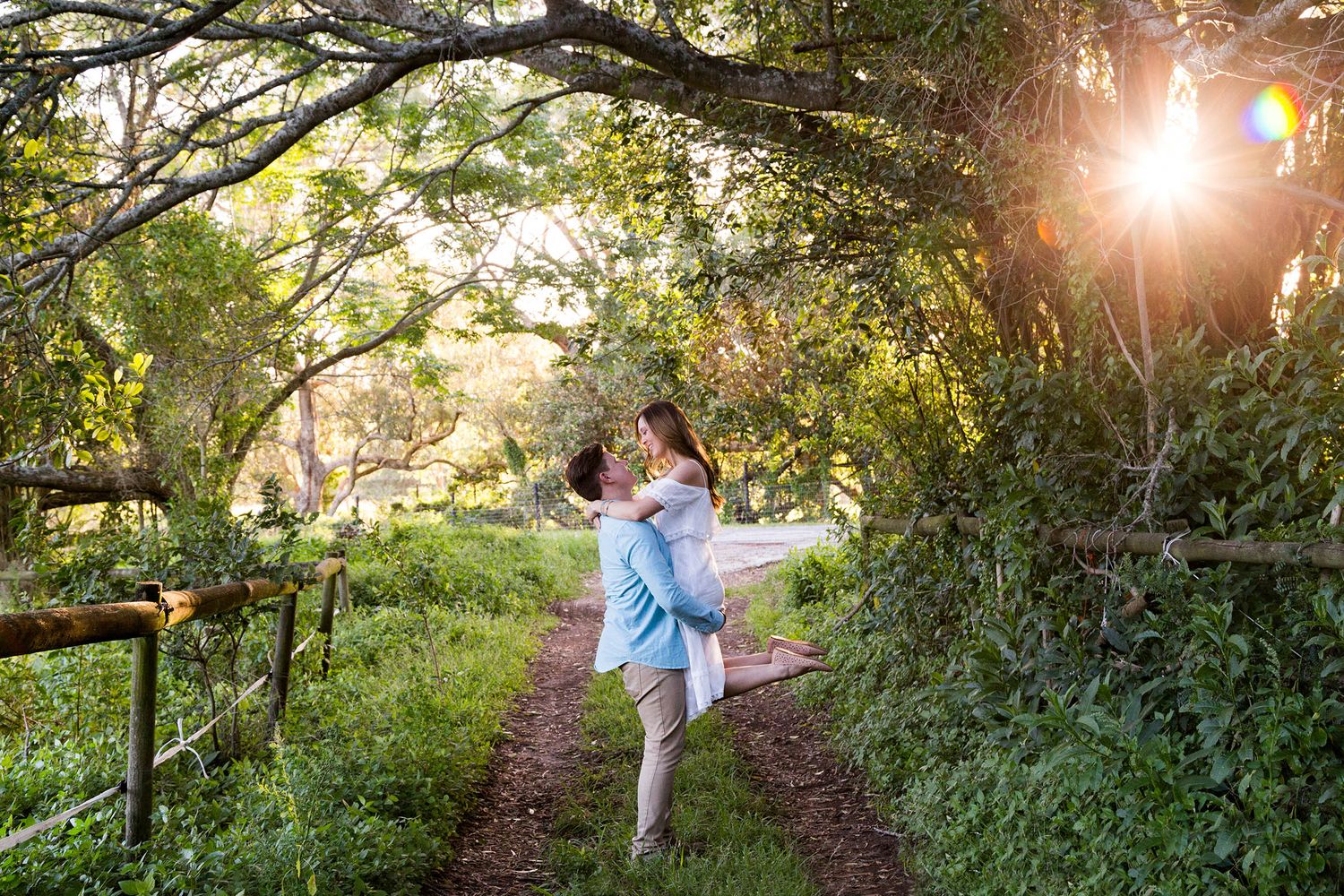 A romantic moment captured on a sunlit garden path beneath overhanging trees during golden hour.