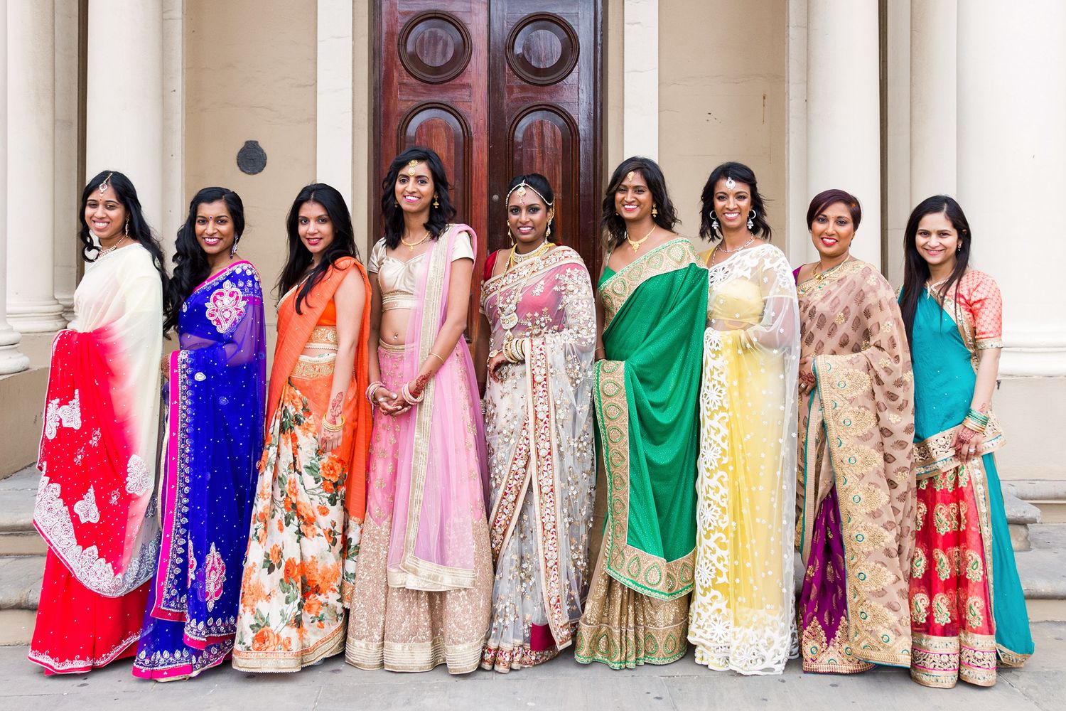 A large group wearing colorful traditional Indian saris and lehengas stands together in an elegant venue.