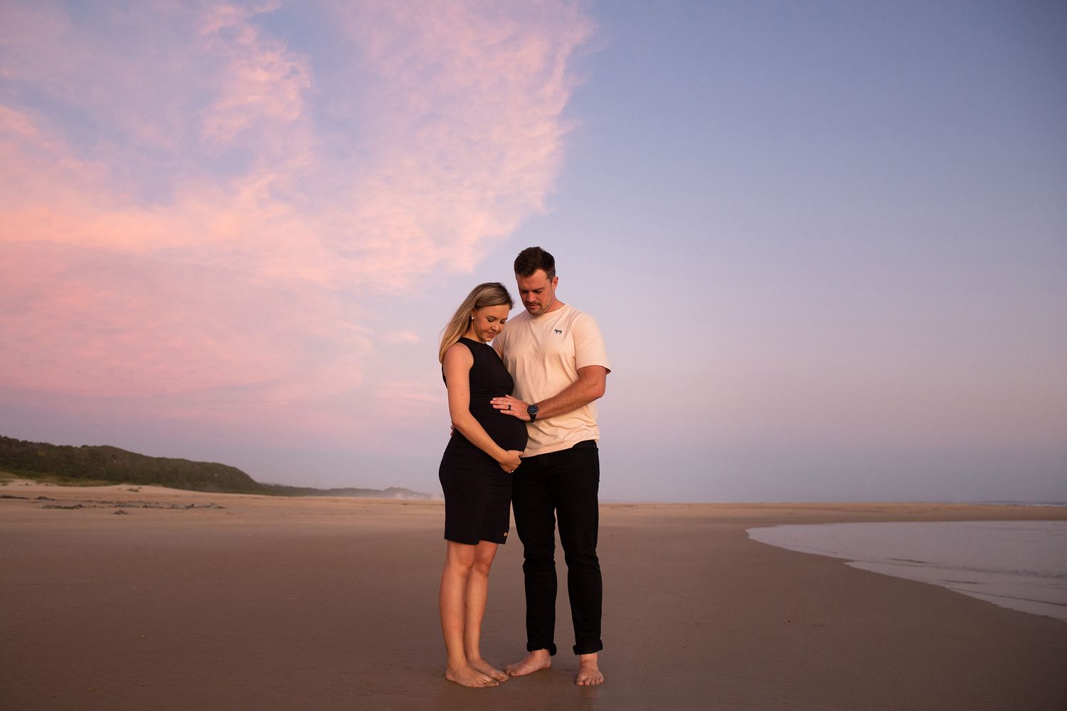 Silhouetted couple stands on beach at sunset with pink and purple sky in background.