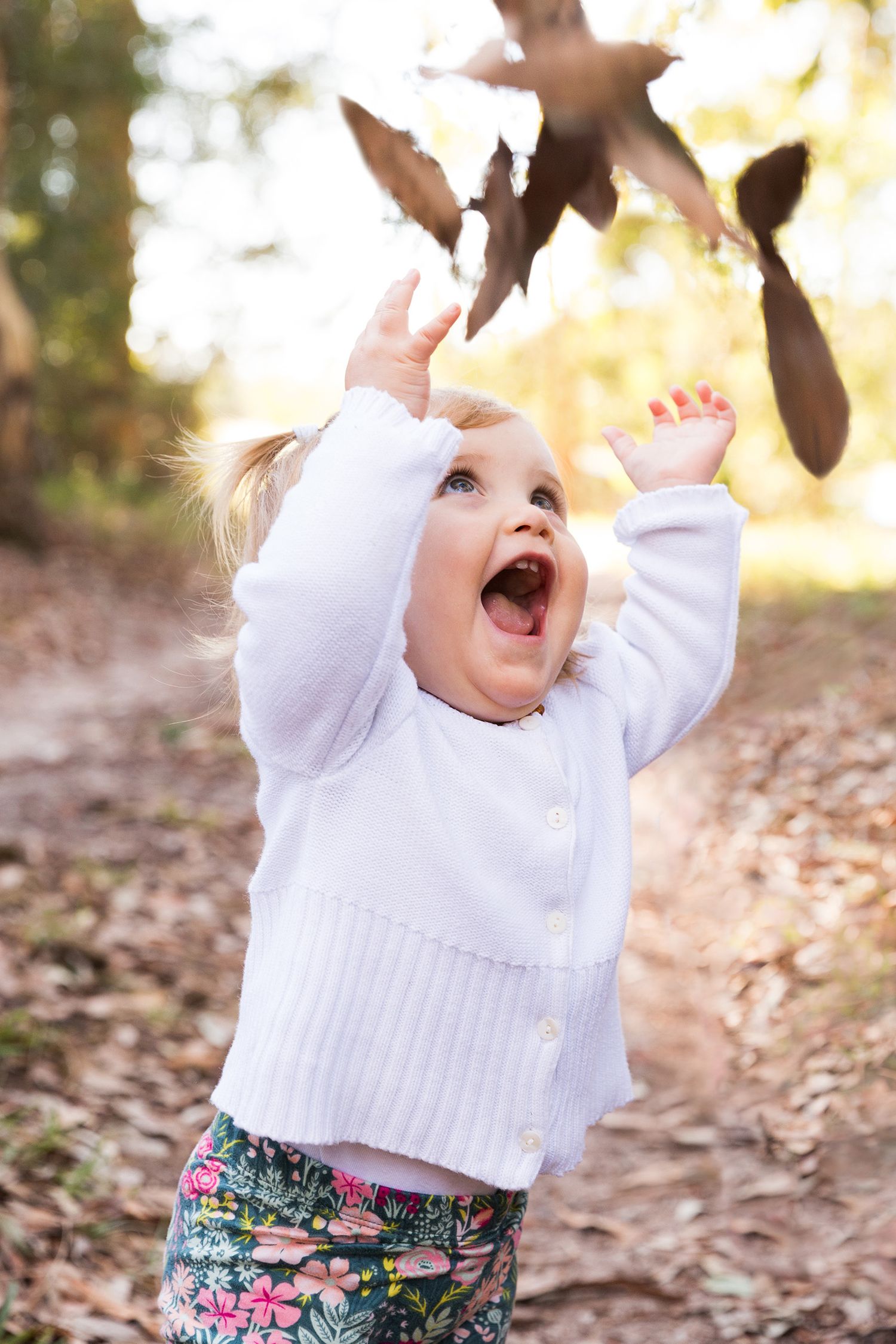 A joyful child in a white sweater plays with falling autumn leaves on a sunlit woodland path.