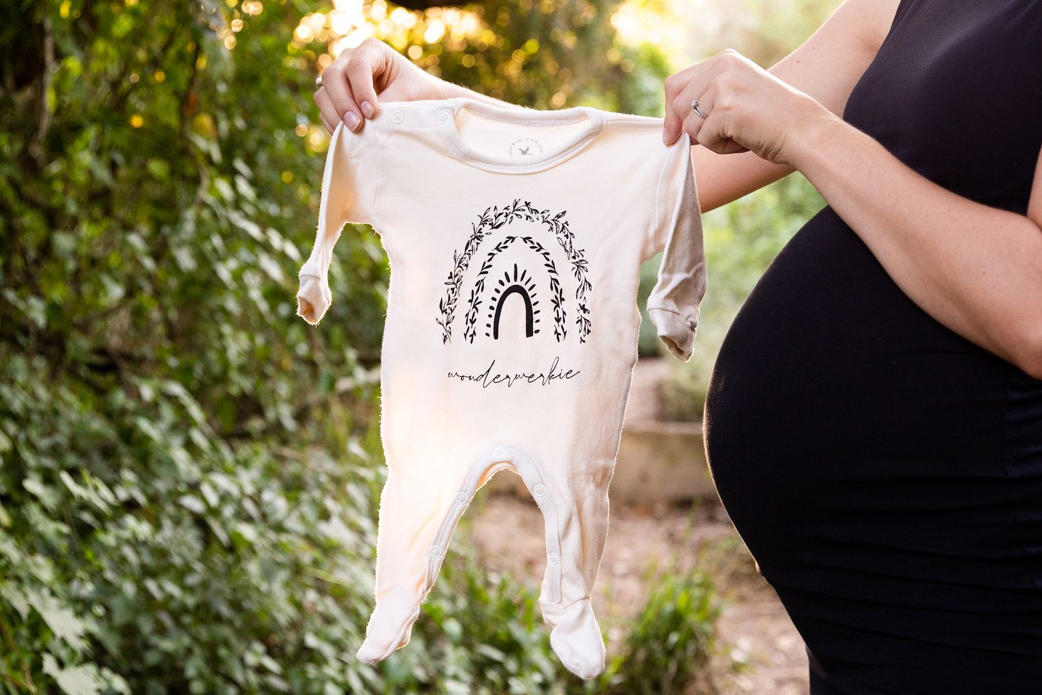 A person wearing black holds up tiny white baby clothing items against sunlit greenery in an outdoor setting.