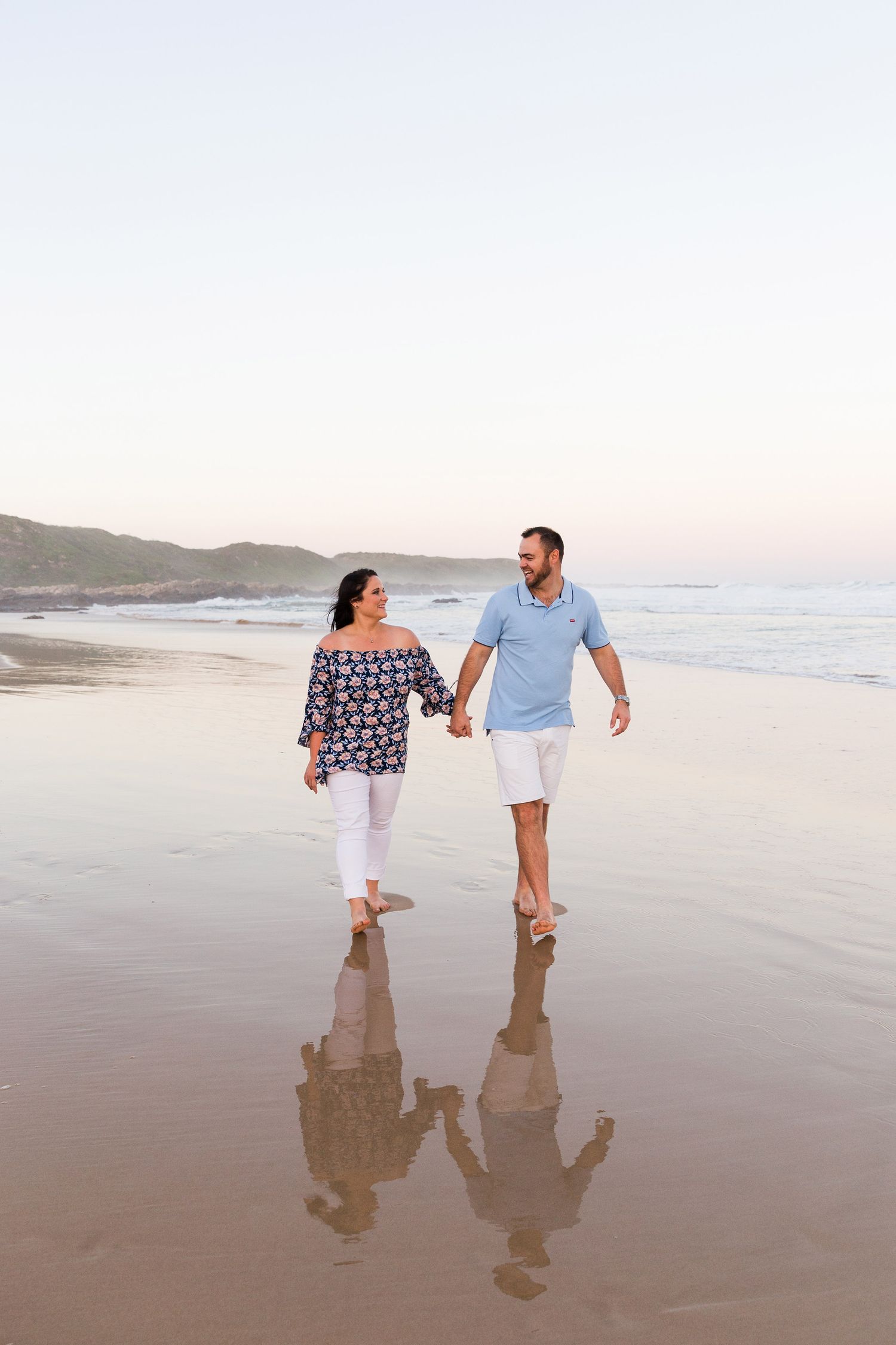 Couple walking along sandy beach at sunset with their reflections visible in wet sand.