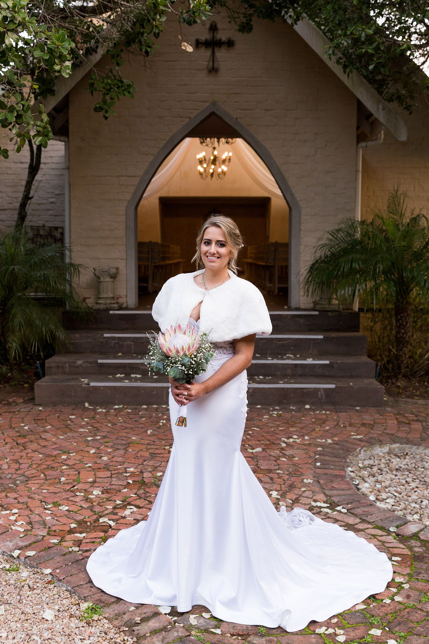 A bride in a white mermaid wedding gown with a short cape stands in front of a rustic chapel entrance.