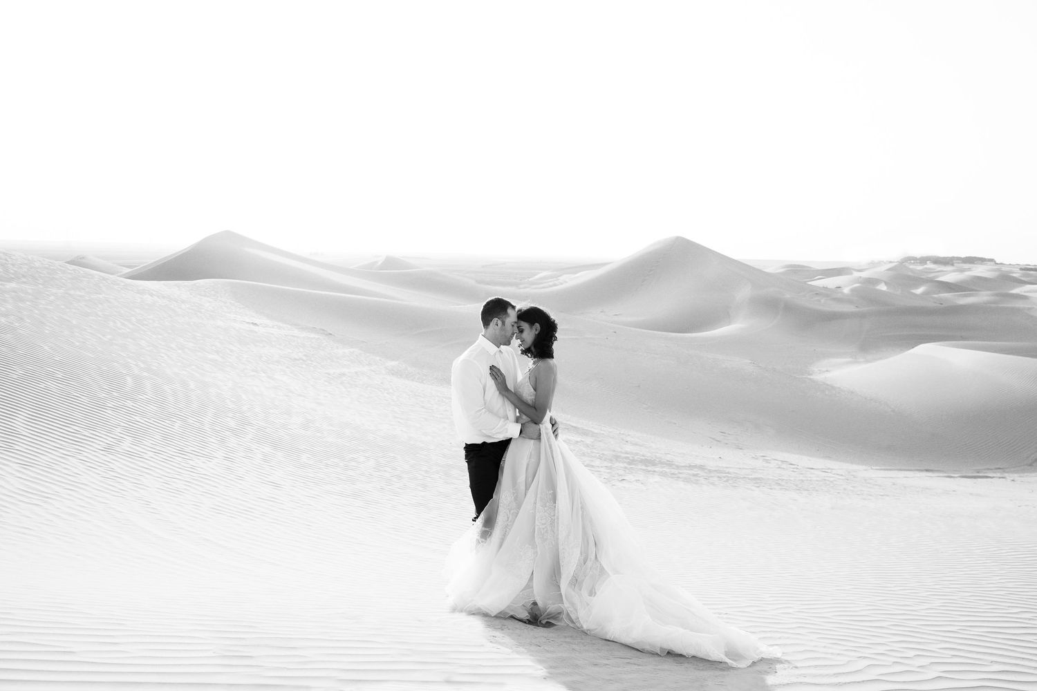 Dramatic black and white photo of a couple in formalwear posing on rolling sand dunes.