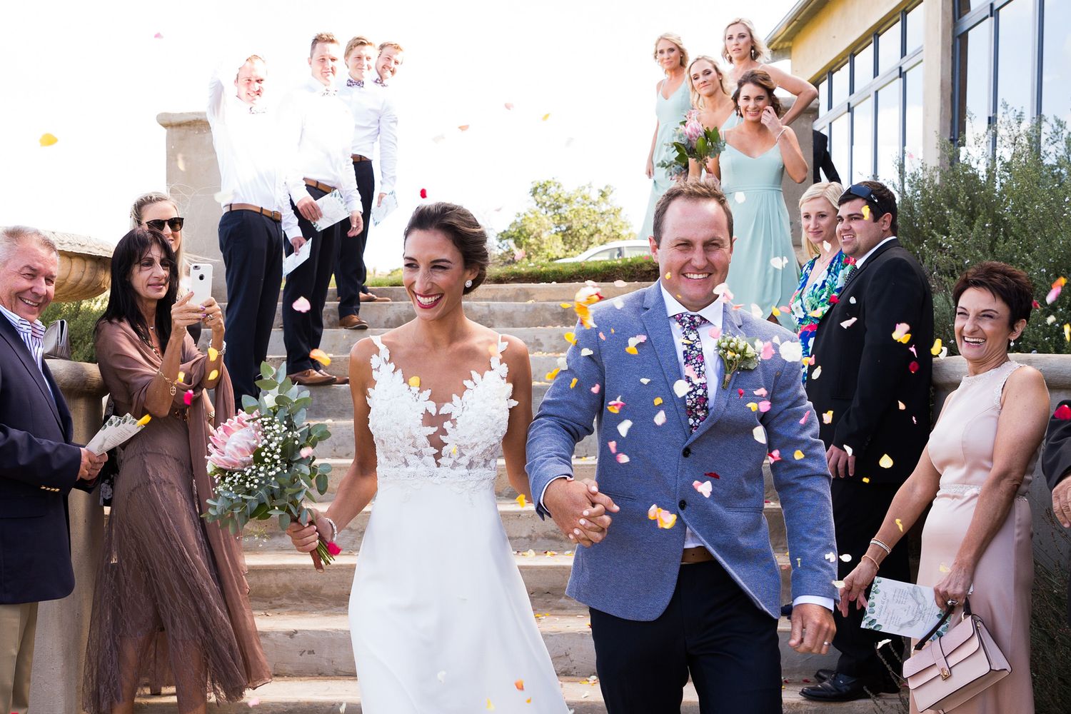 Wedding guests throw confetti as they celebrate at an outdoor reception on a sunny day.