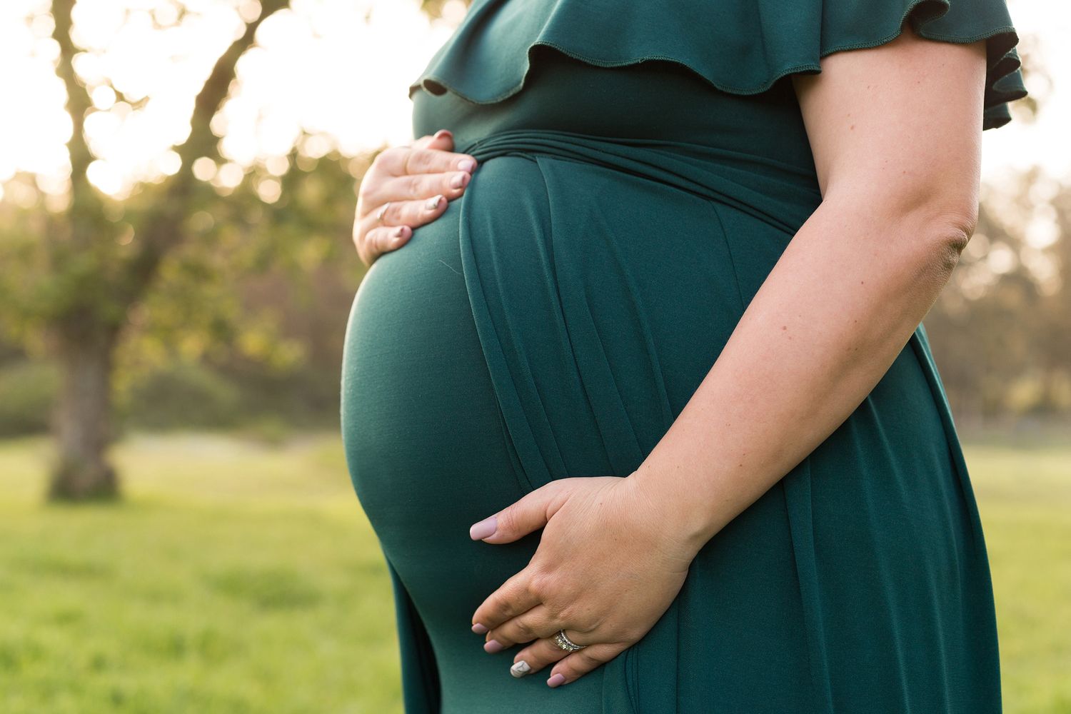 A person wearing a long green maternity dress stands in a field during golden hour.
