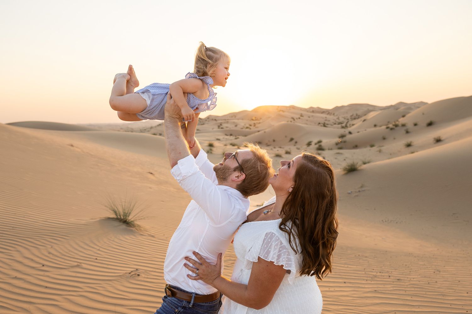 Two people embrace on desert sand dunes at sunset with warm golden light casting long shadows across the landscape.