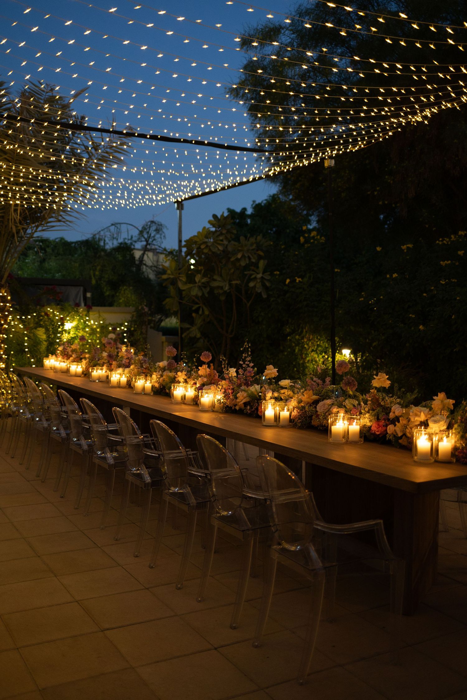 Outdoor evening wedding reception setup with string lights and lantern centerpieces on long tables.