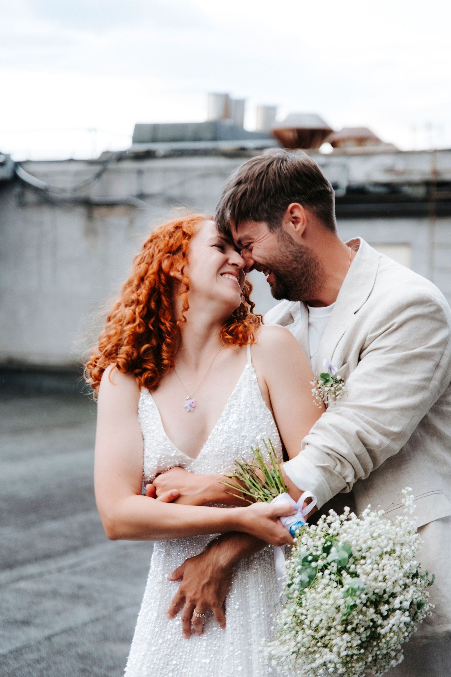 Romantic wedding couple embraces while holding delicate baby's breath bouquet on rooftop.