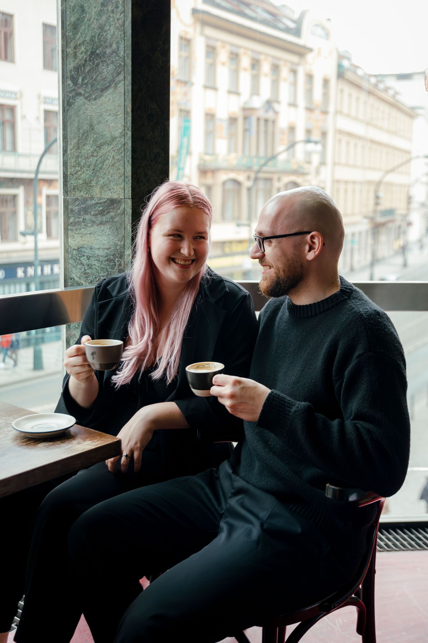Couple shares lighthearted moment while having coffee at window-side cafe table.