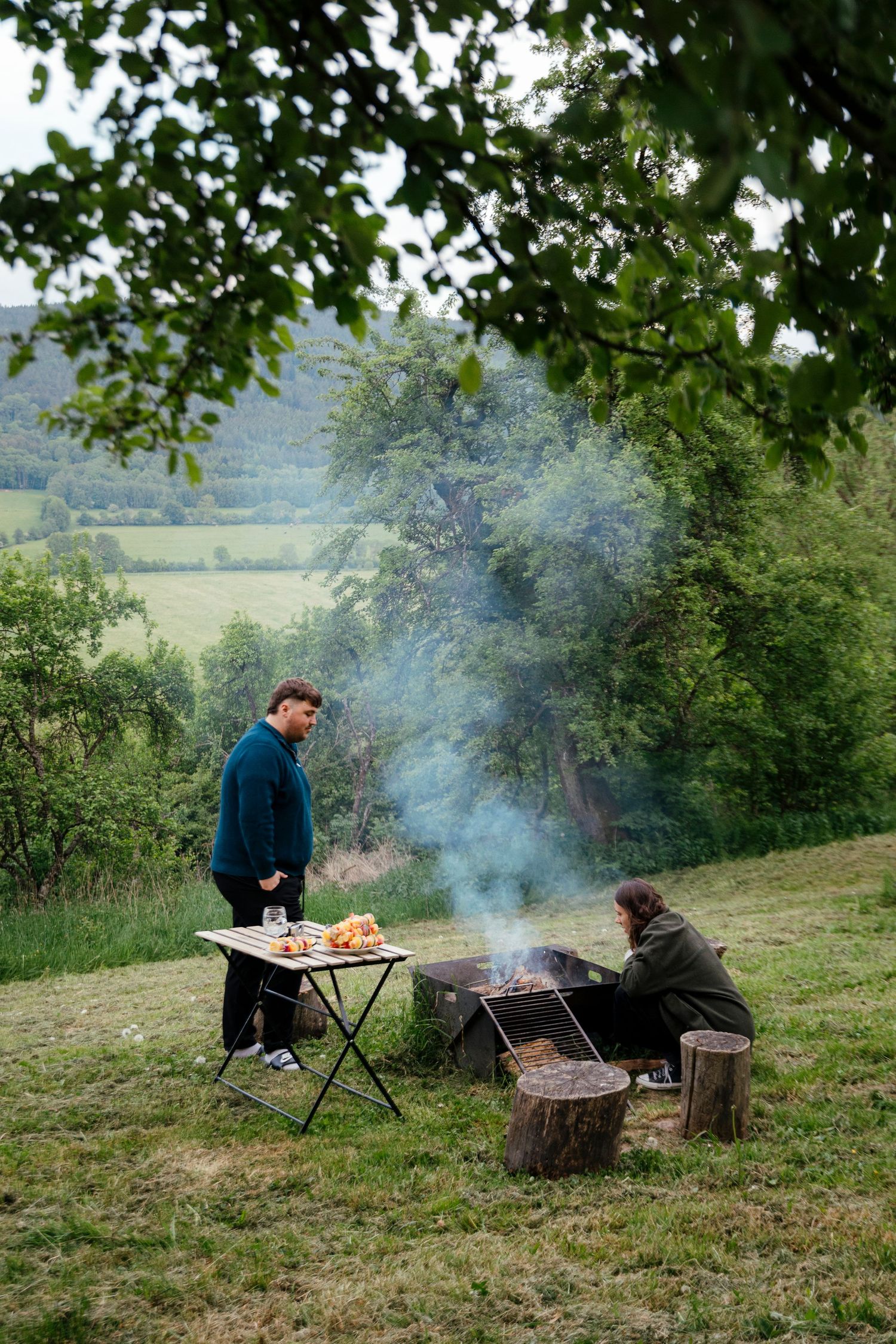 Two people grilling food outdoors on a portable barbecue with smoke rising through the trees.
