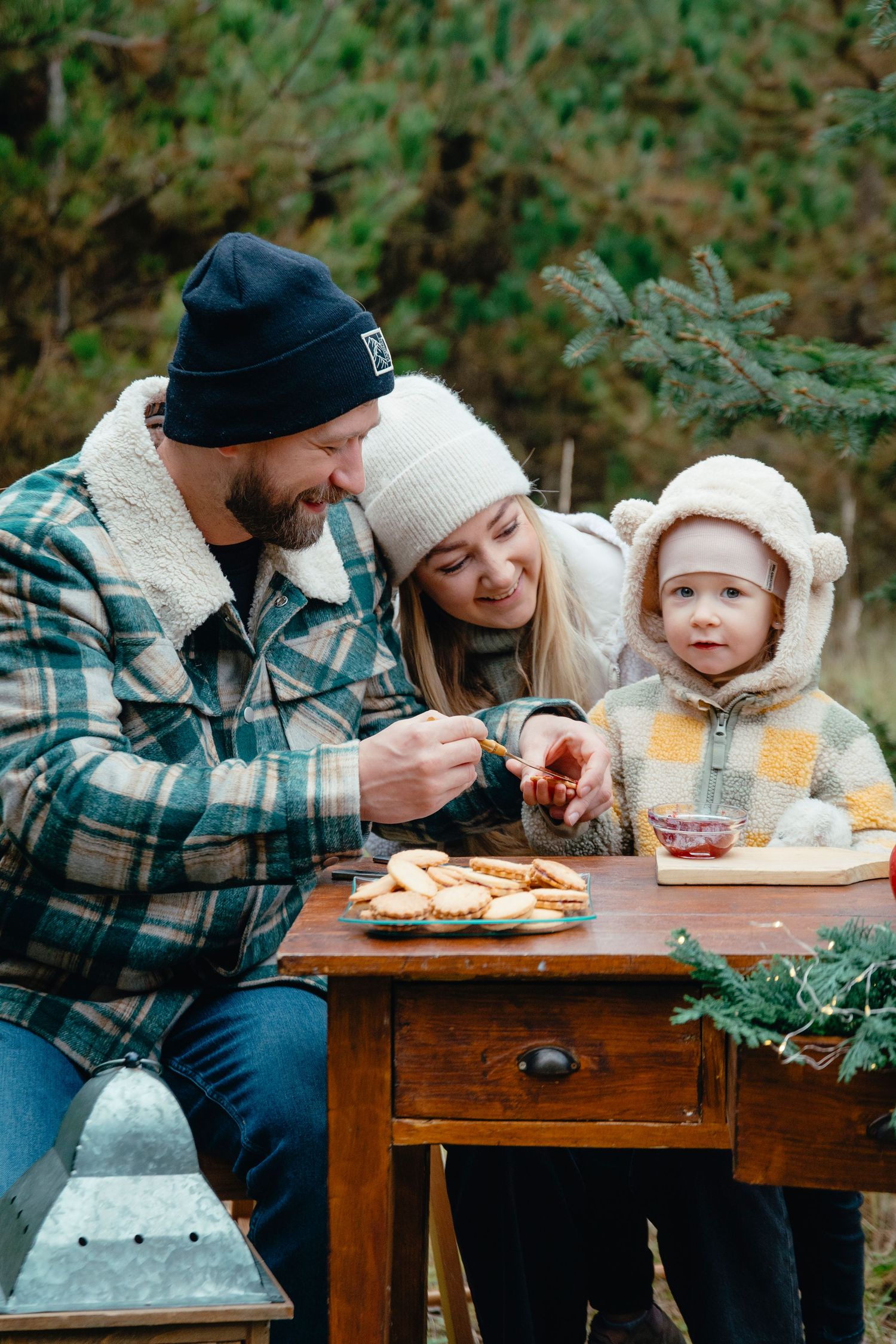 Family with toddler enjoying outdoor winter picnic with cookies and jam at rustic wooden table surrounded by evergreen trees.