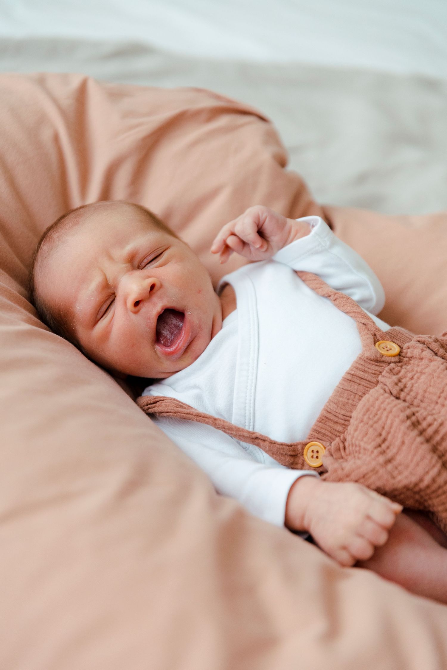 Newborn baby yawning while lying on pink bedding, dressed in white onesie and brown linen overalls.